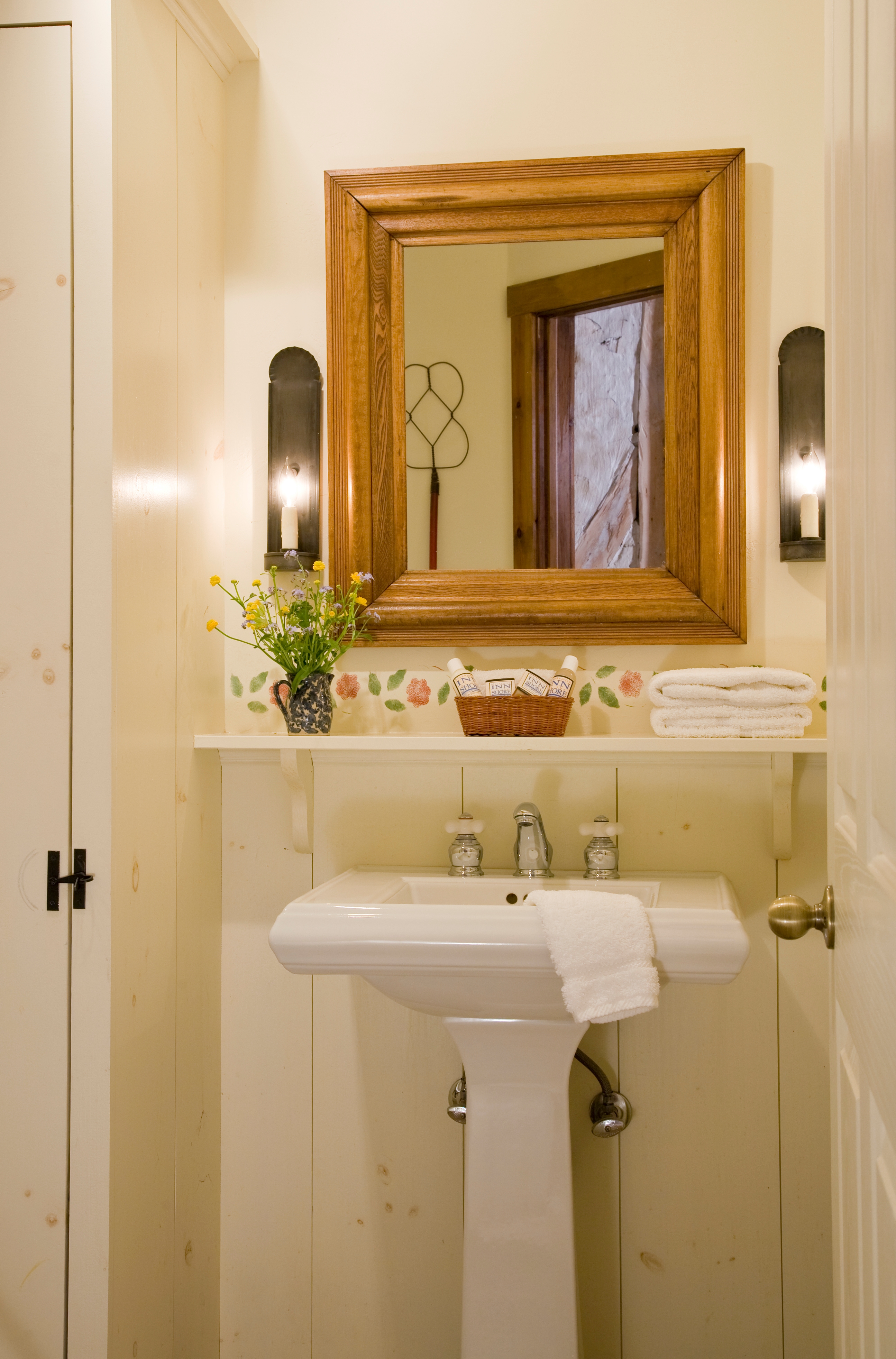 A bathroom vanity featuring a white pedestal sink, a wood-framed mirror flanked by two wall sconces, and a small shelf holding flowers and folded towels.