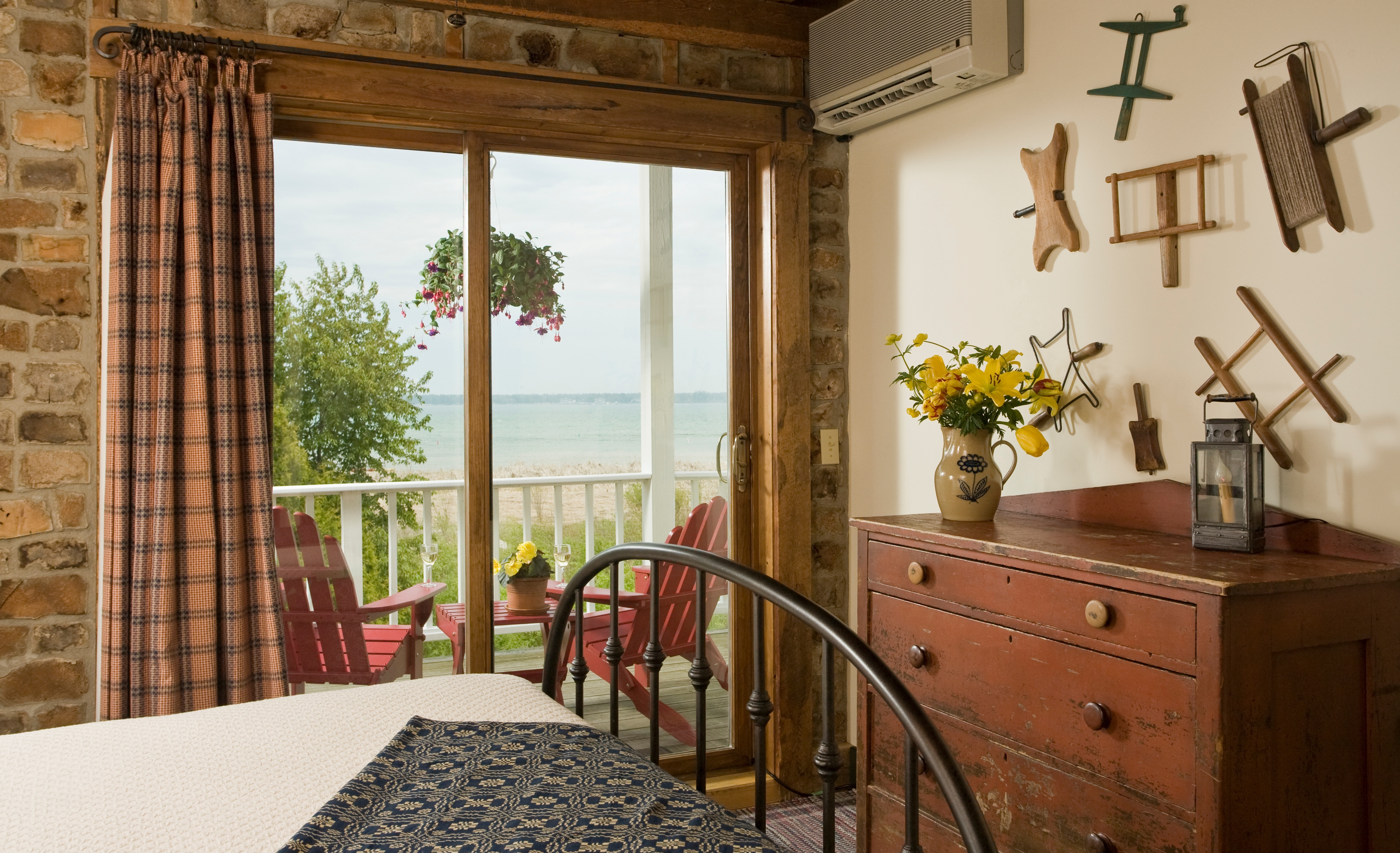 A bedroom interior featuring a wooden dresser with a vase of yellow flowers and a sliding glass door that opens to a balcony with red chairs and a view of the harbor.
