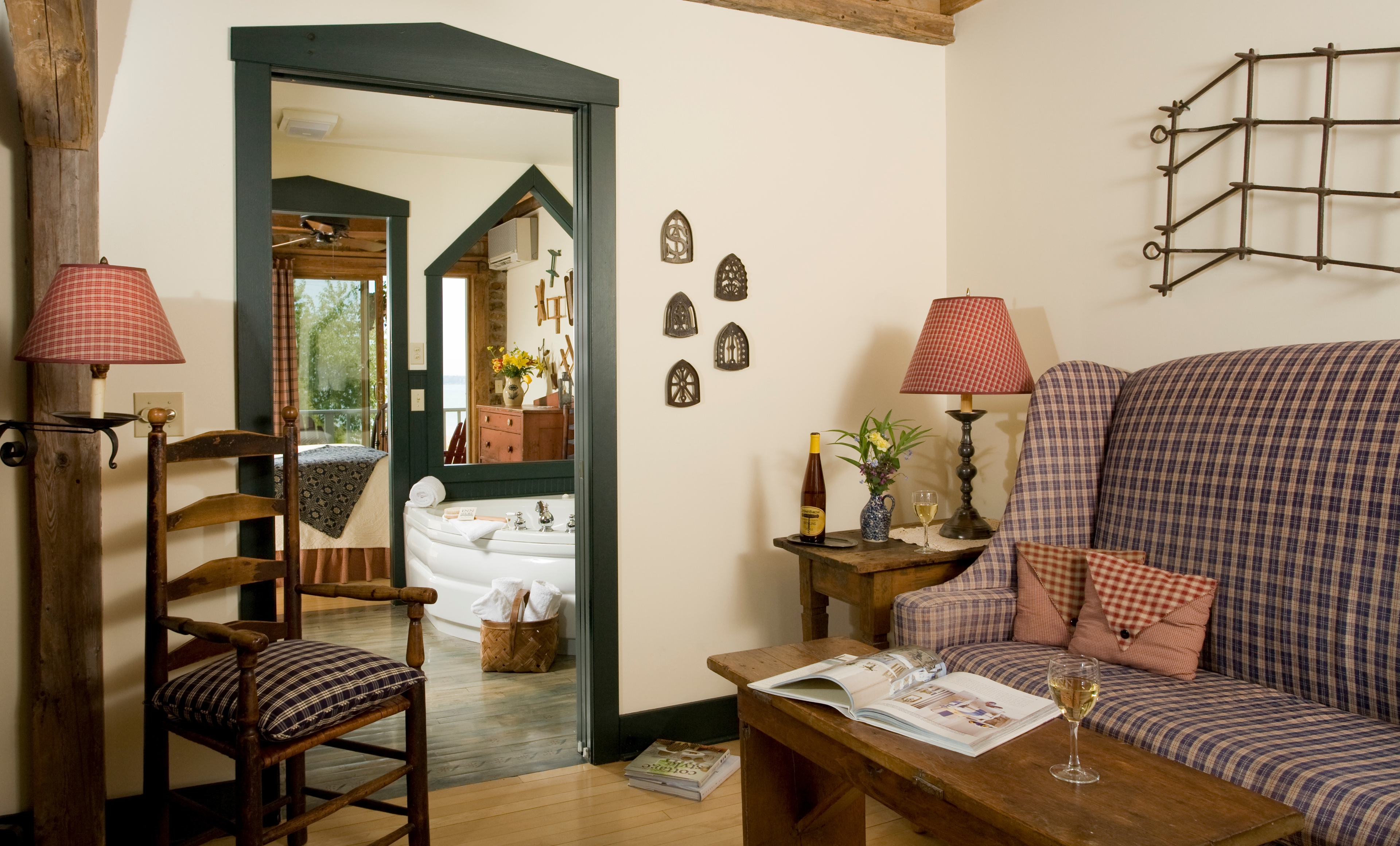 View through a dark green doorway into a bathroom featuring a large white whirlpool tub, with a wooden ladder-back chair in the foreground and a reflection in the mirror showing a bedroom with a view of the water.