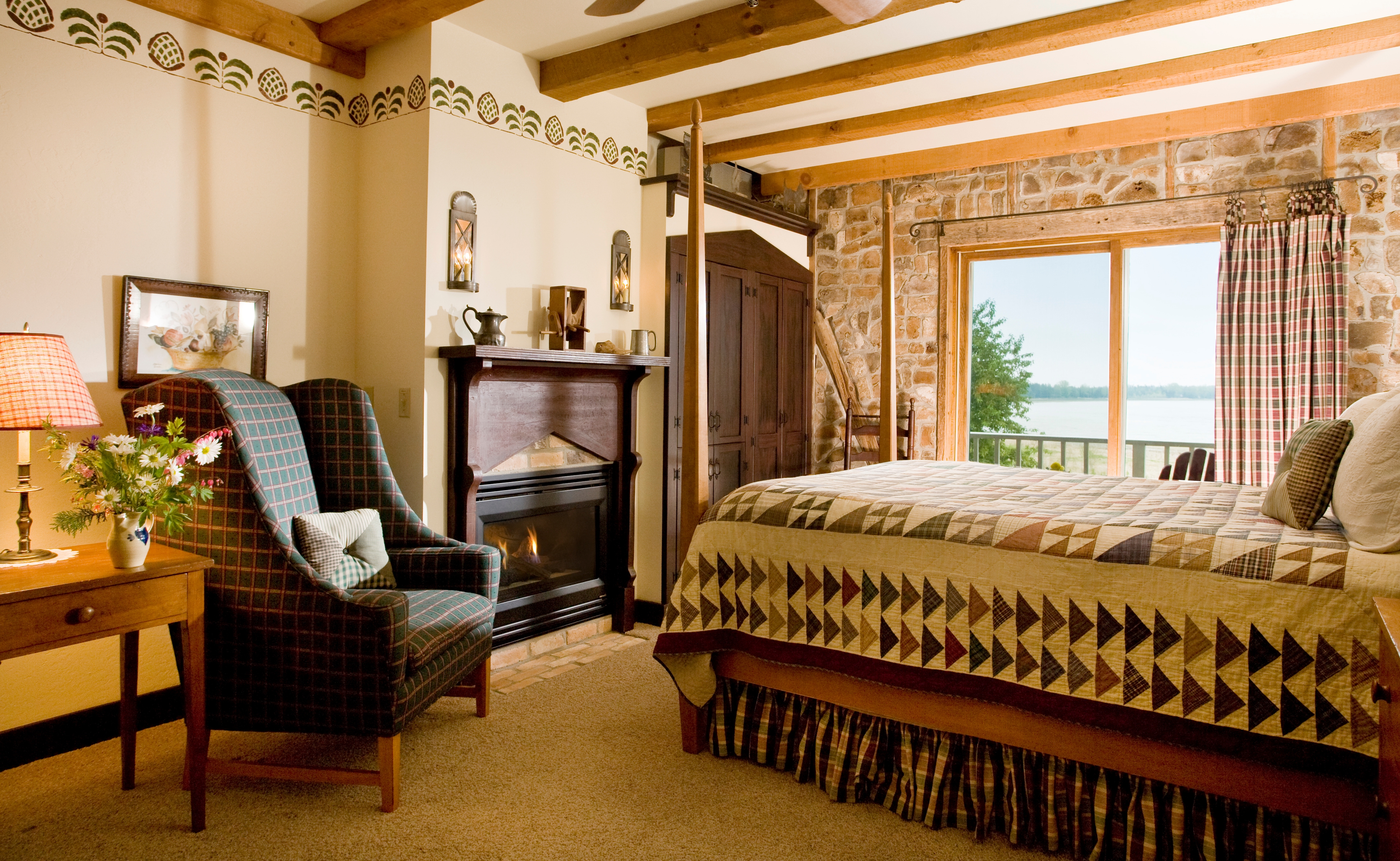A cozy bedroom featuring a patterned quilt on a bed, a high-back armchair next to a fireplace, and a sliding glass door that offers a scenic view of Lake Michigan.