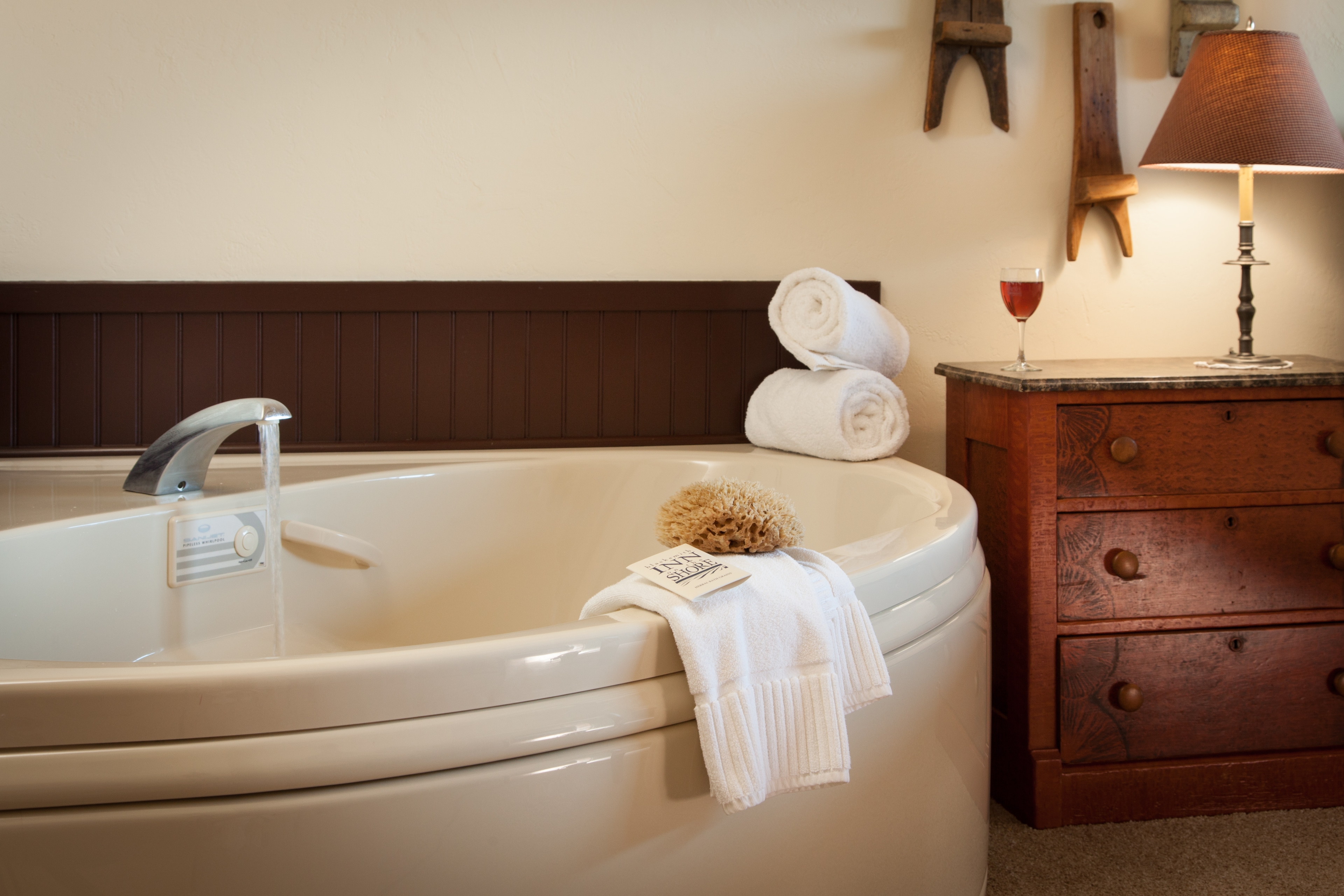 A bedroom corner featuring a wooden three-drawer nightstand with a lit lamp and a glass of wine, positioned between a large white whirlpool tub and a black metal canopy bed.