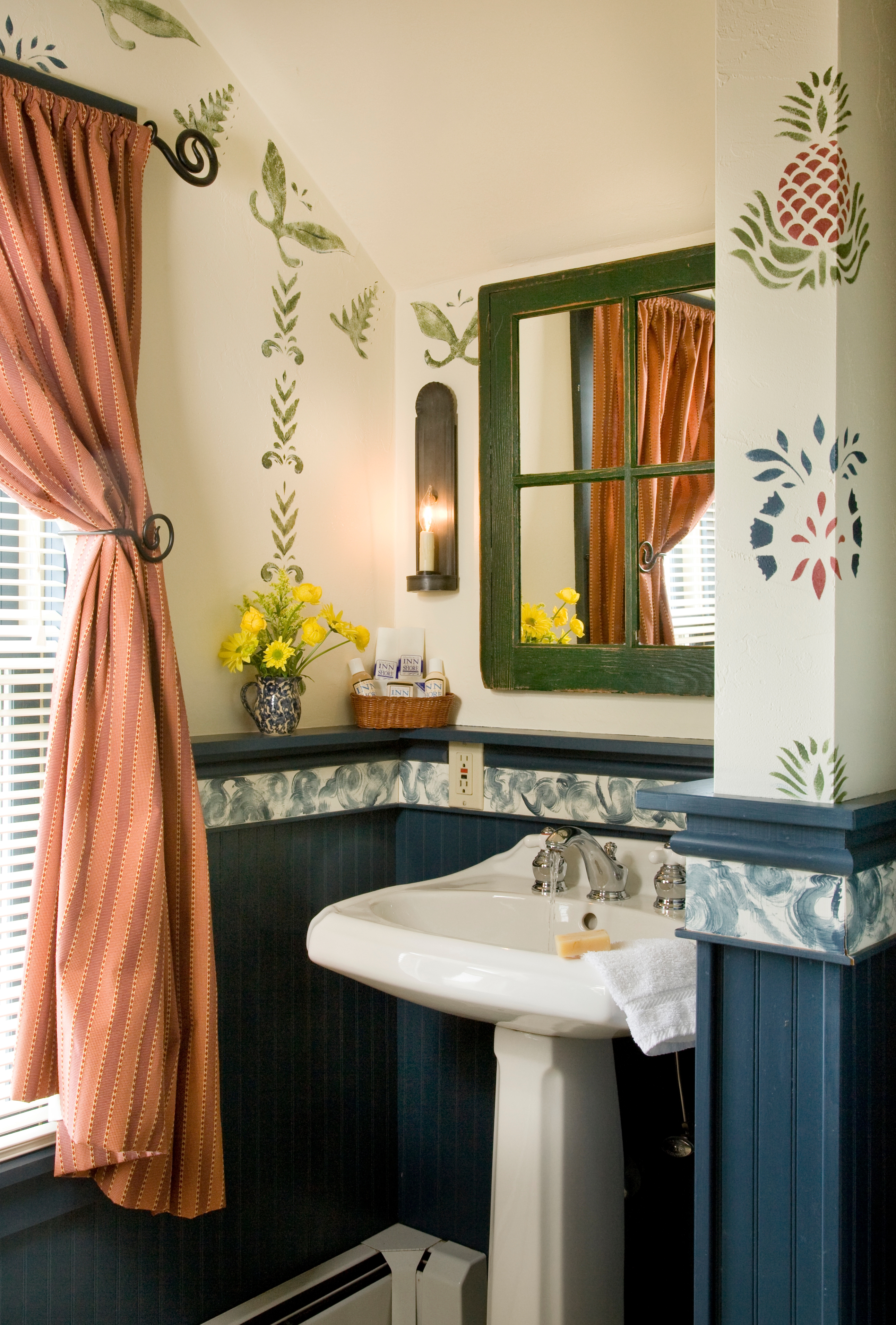 A white pedestal sink in a bathroom featuring blue wainscoting and cream-colored walls decorated with folk-art stencils of pineapples and vines, with a green-framed mirror and a small vase of yellow flowers on a shelf.