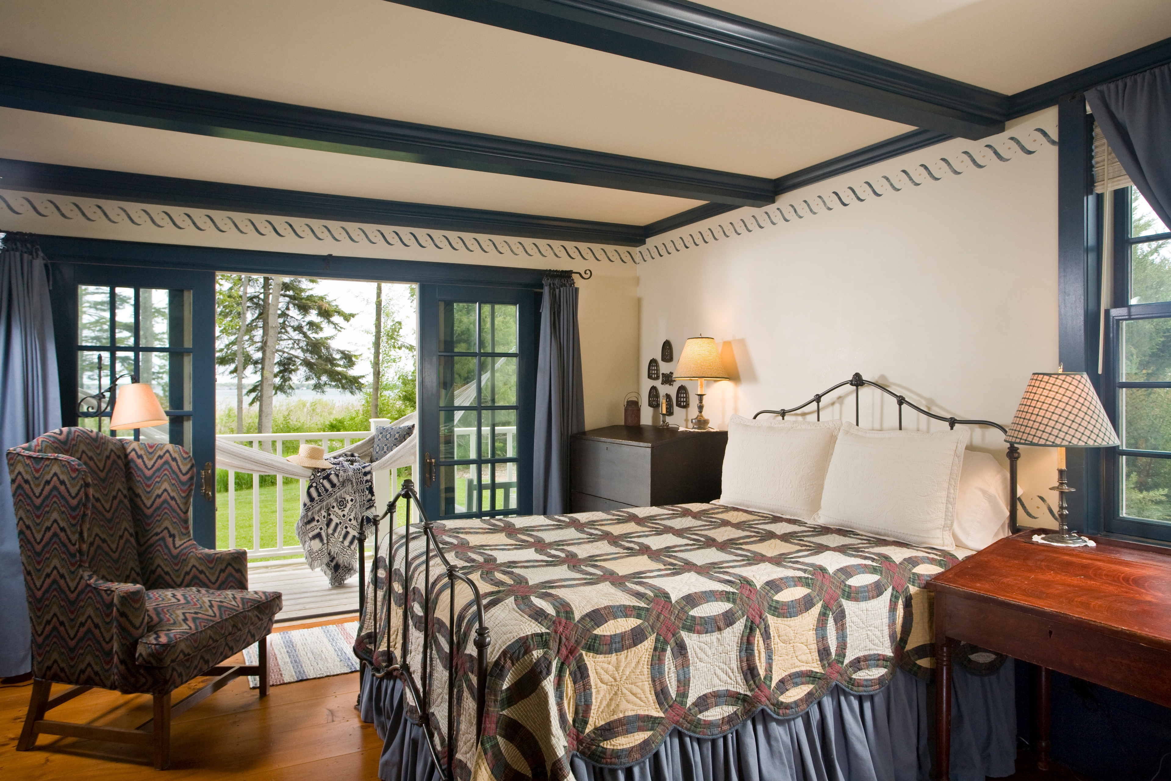 A bedroom with blue-painted ceiling beams and trim, featuring a bed with a double wedding ring patterned quilt, a patterned wingback chair, and french doors that open to a balcony with a hammock and a view of trees.