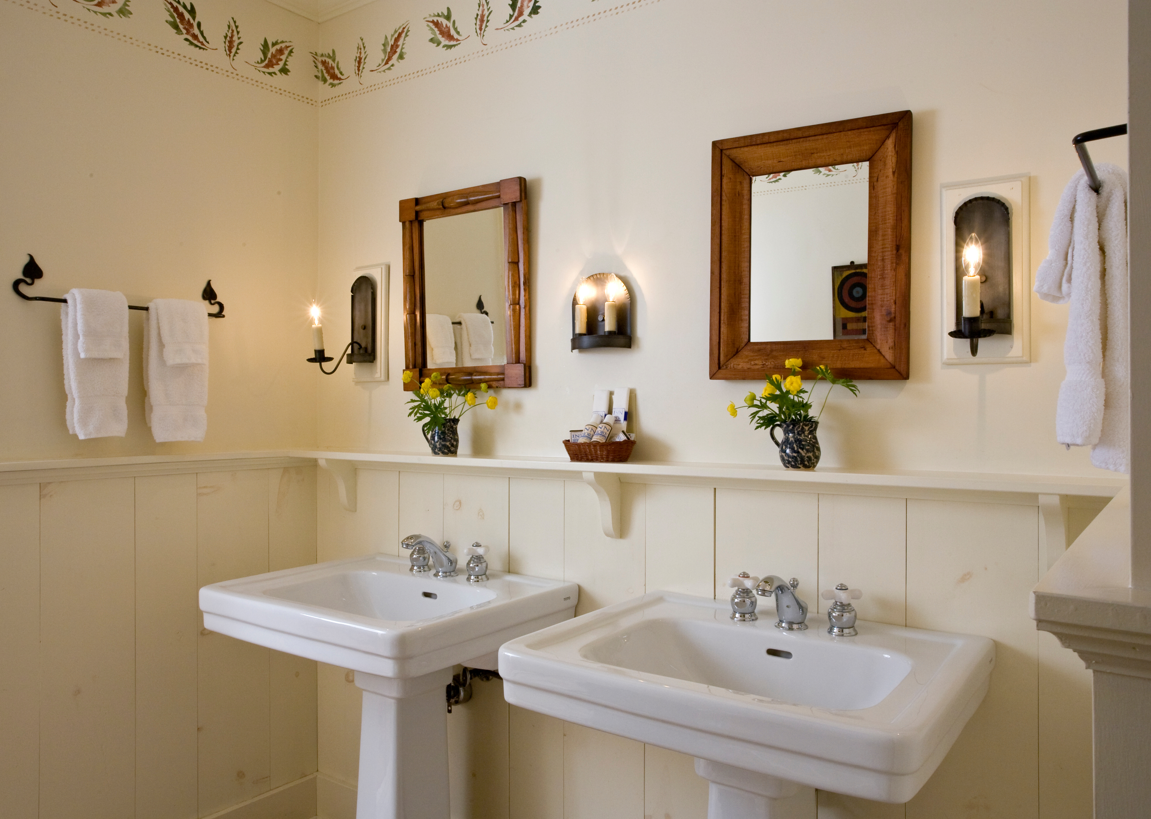 A bathroom featuring two side-by-side white pedestal sinks, each with its own wood-framed mirror and a small vase of yellow flowers on a shelf above.