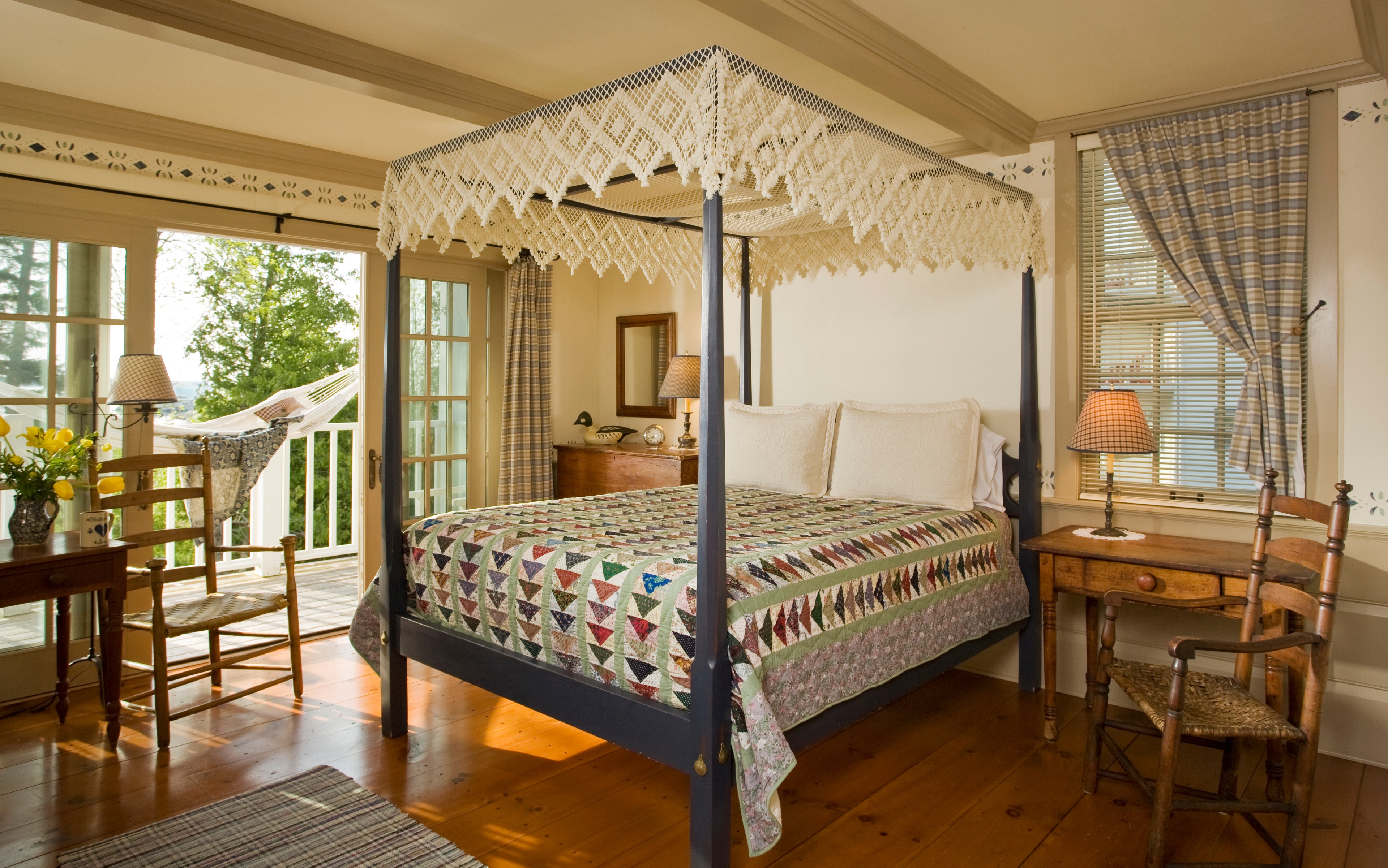 A spacious bedroom featuring a black canopy bed with a white macramé canopy and a colorful geometric patterned quilt, a wooden desk with a lamp, and French doors leading to a balcony with a white hammock and a view of greenery.