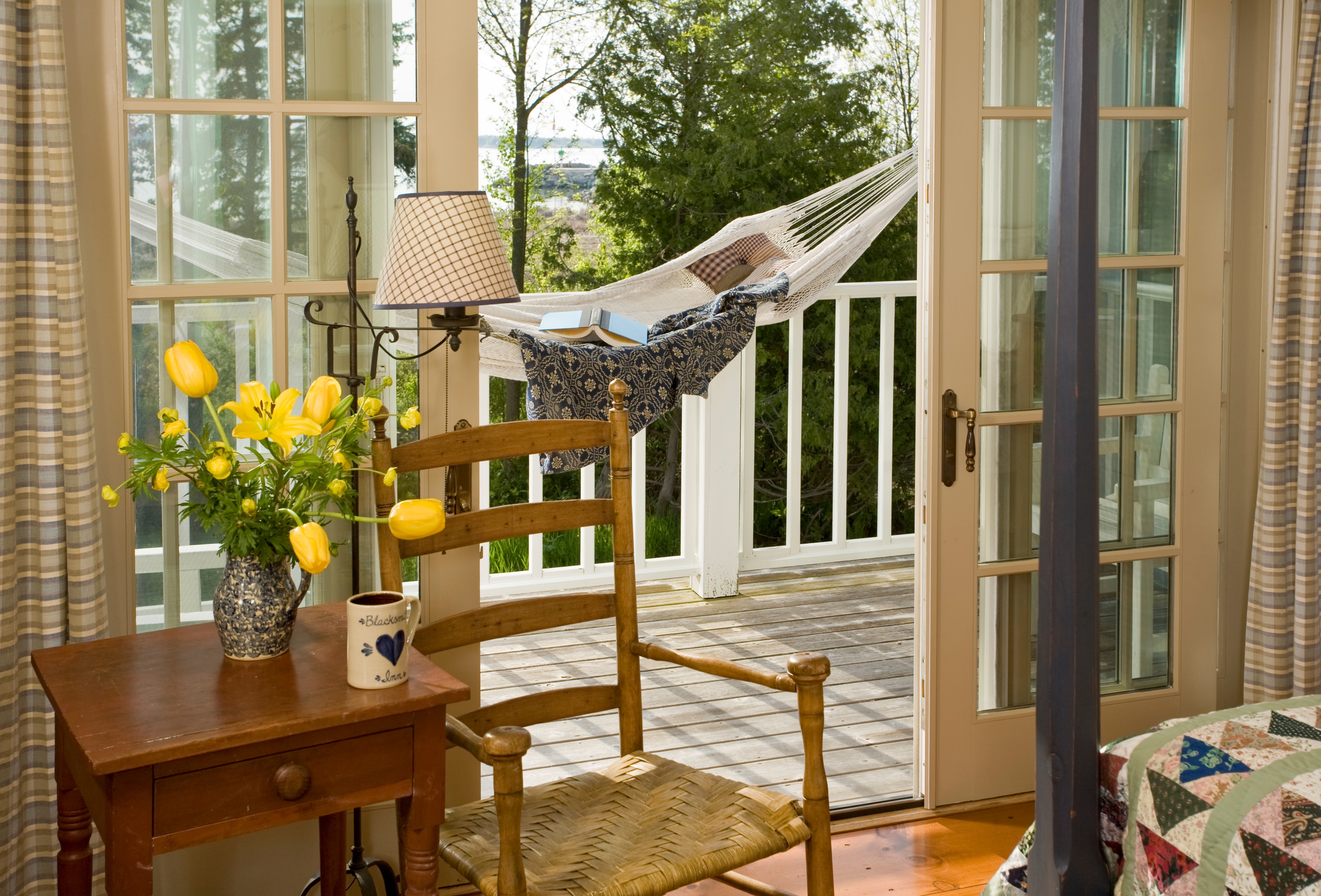 A cozy bedroom corner featuring a wooden ladder-back chair and a small side table with a mug and a vase of yellow flowers, set next to an open door that leads to a sunlit balcony with a white hammock.