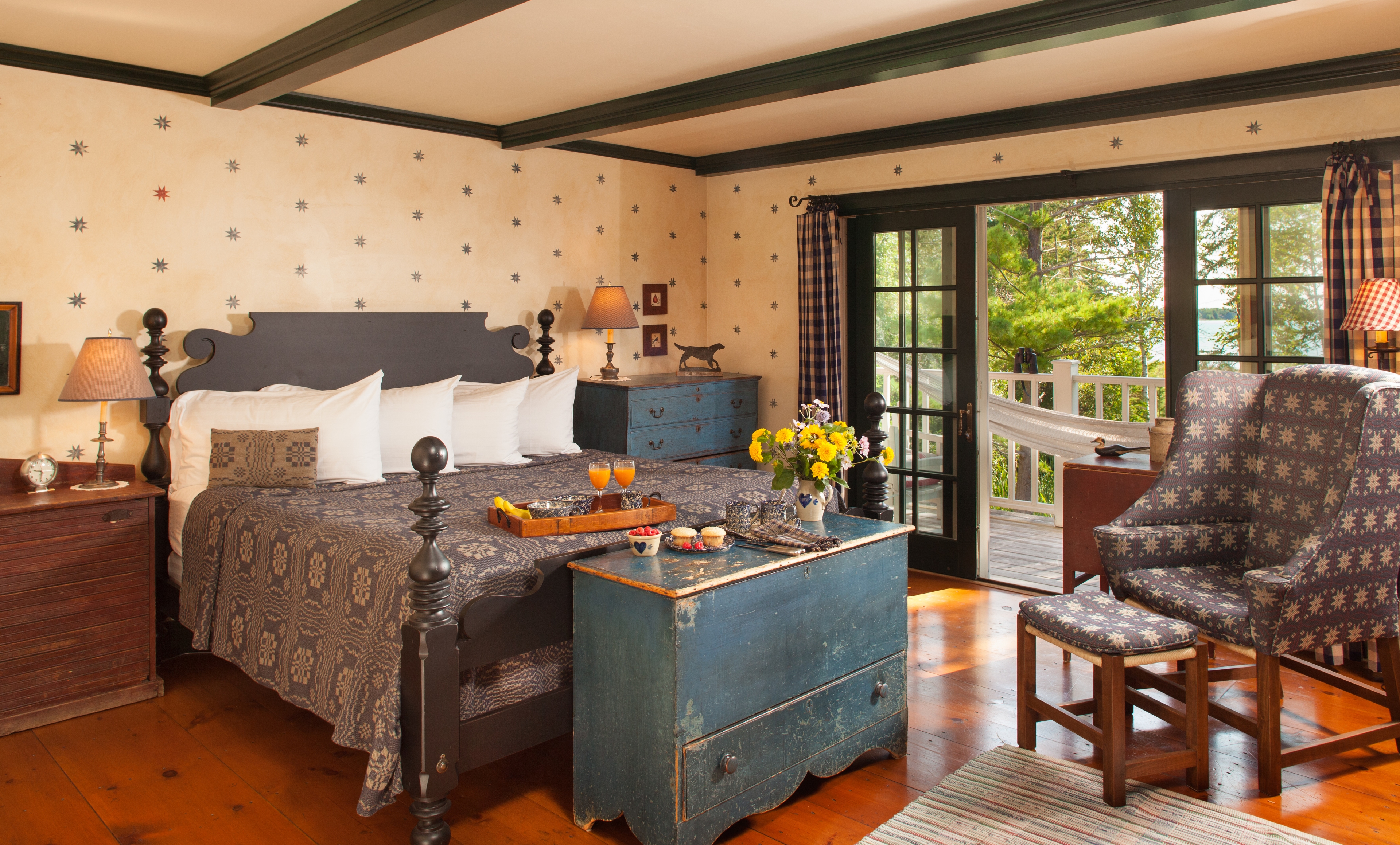 A spacious bedroom with a dark wood bed and a gray patterned quilt, featuring a blue chest at the foot of the bed and a matching blue dresser. Large glass doors lead to a sunlit balcony with a white hammock and a view of lush green trees.