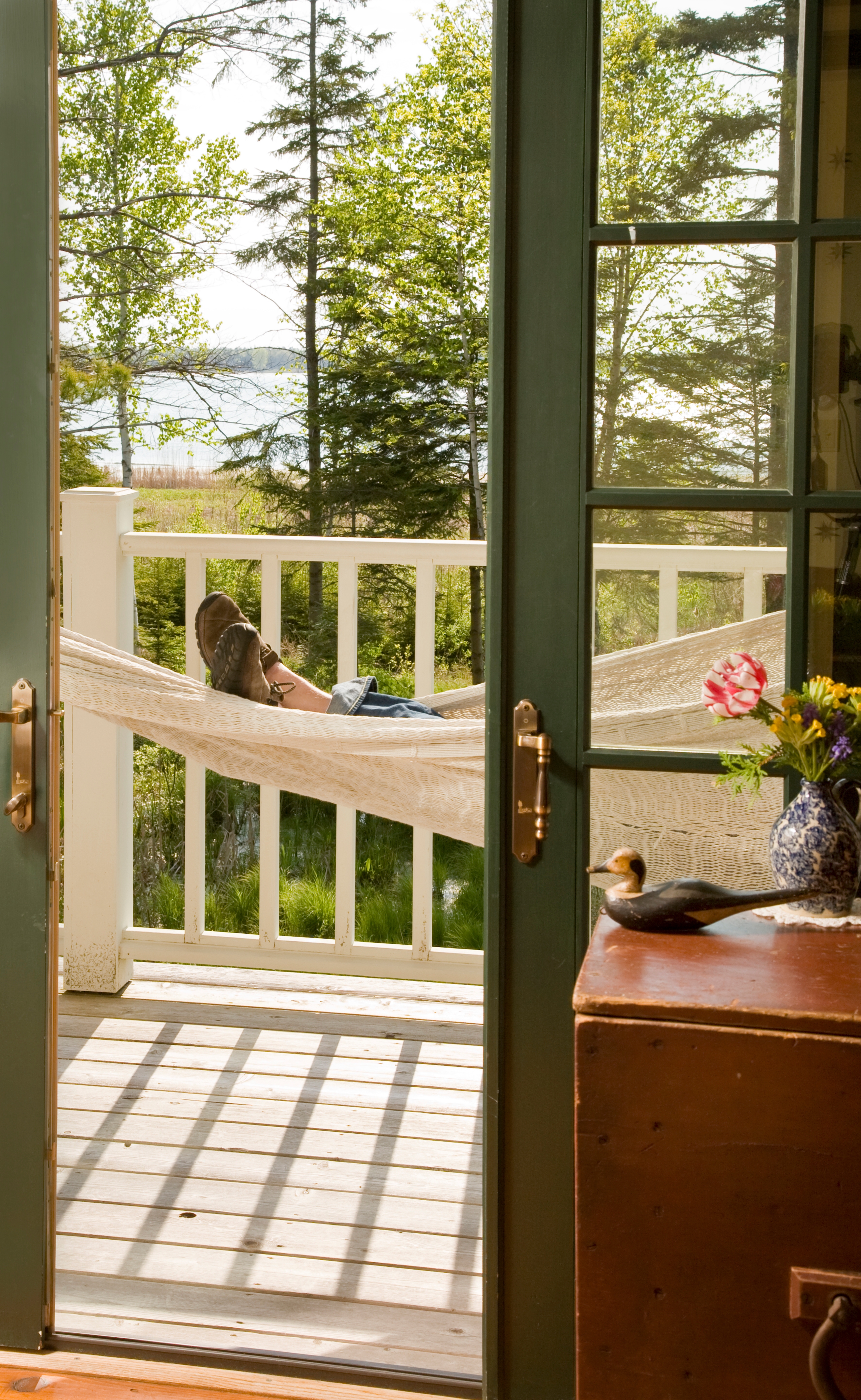 A close-up view of a person's feet resting in a white rope hammock on a balcony, with a white railing and a scenic background of green trees and a body of water.