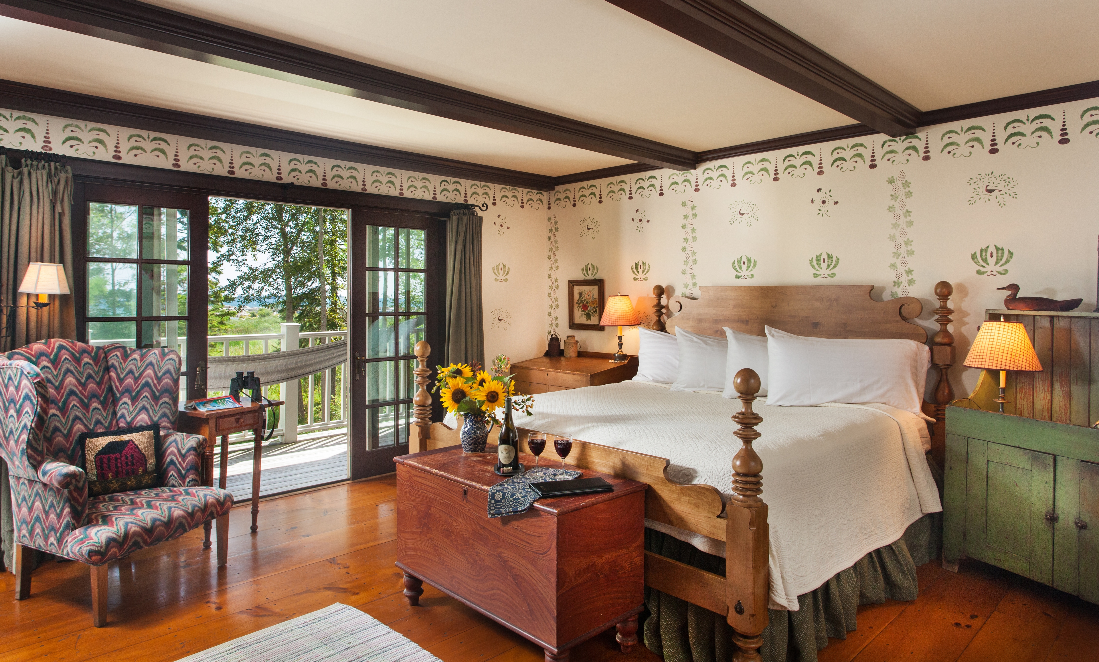 A spacious bedroom with white-stenciled walls and dark ceiling beams, featuring a large wooden four-poster bed with white linens, a wooden chest with sunflowers and a bottle of wine at its foot, and a patterned wingback chair. Large glass doors open to a balcony with a white hammock and a view of lush trees and lake shore.
