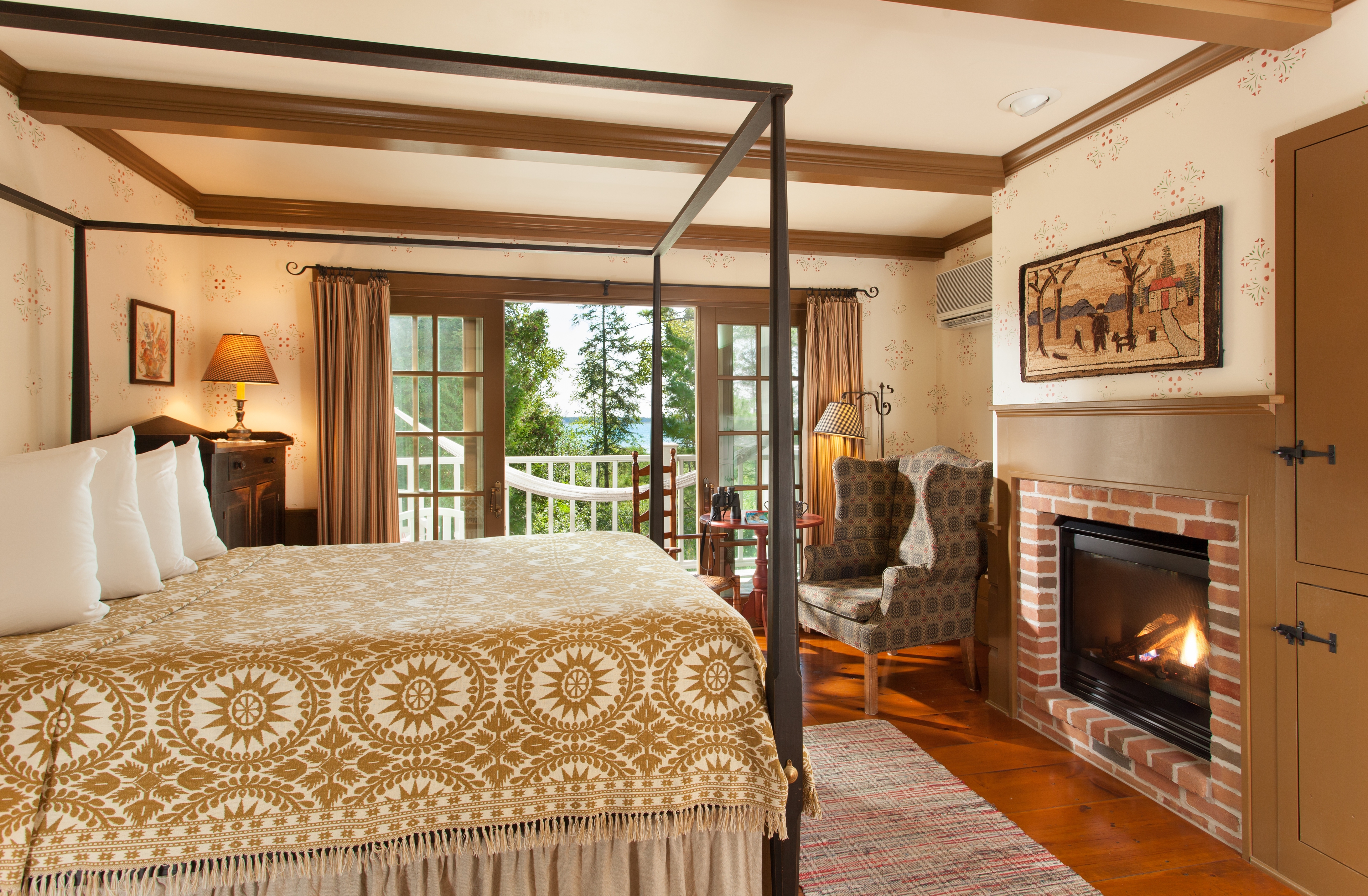 A spacious bedroom featuring a black metal canopy bed with a tan and white patterned quilt, a brick fireplace with a roaring fire, and glass doors leading to a balcony with a white hammock and a view of lush greenery.