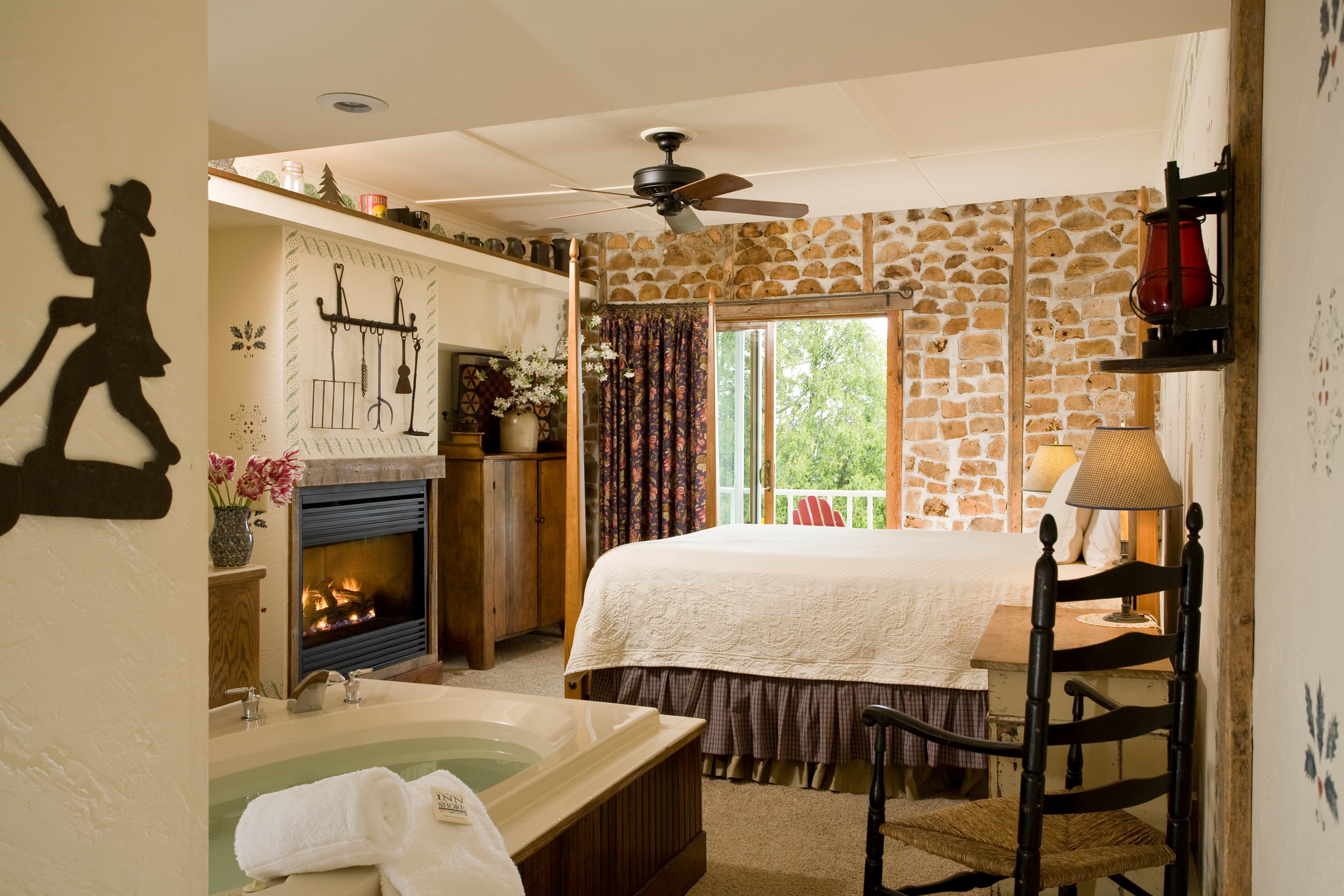 A view from a bathroom featuring a whirlpool tub into an adjoining bedroom with a lit fireplace, a bed with a white comforter, and a stone accent wall next to a balcony door.