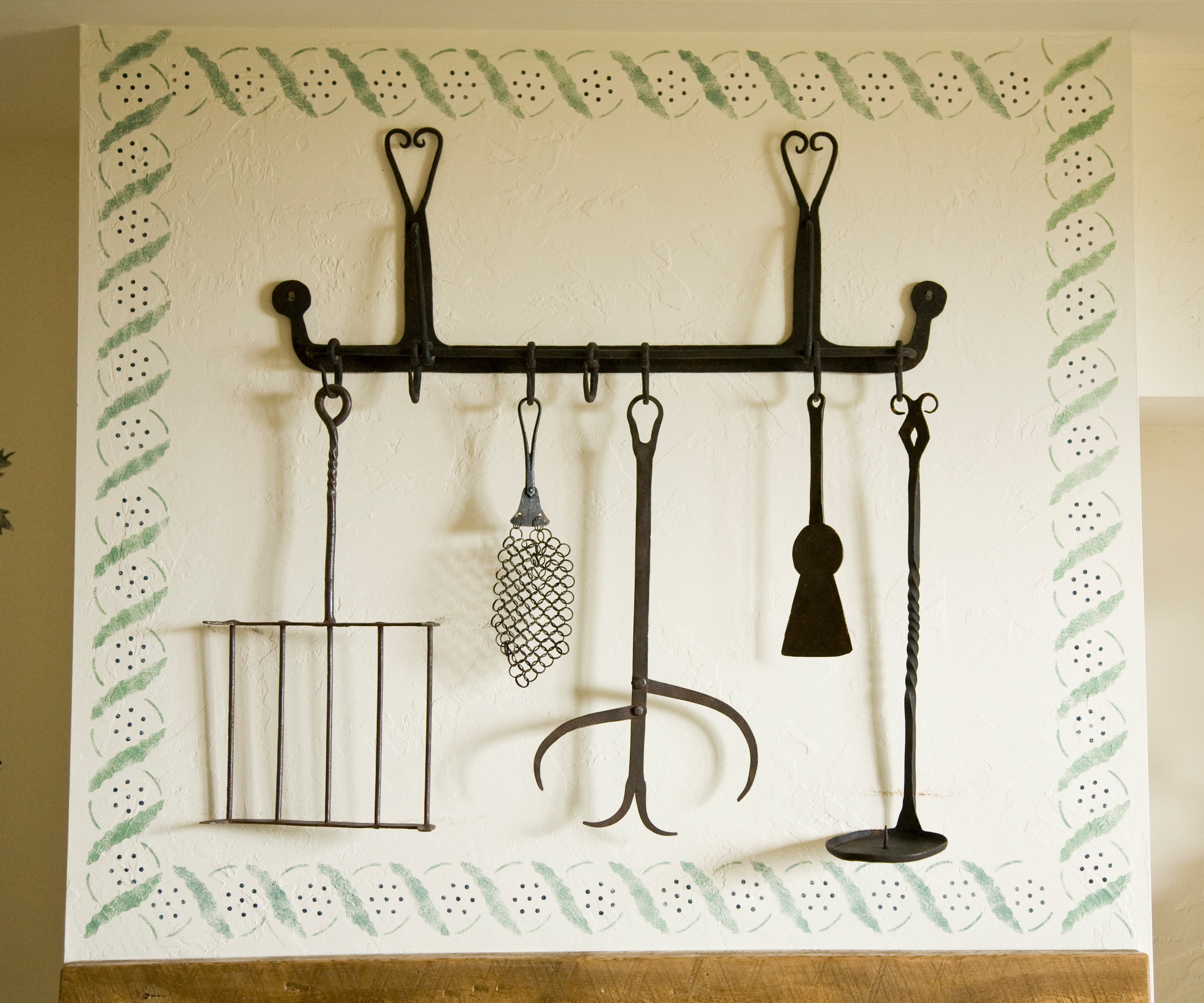 A collection of antique wrought-iron hearth tools, including a toaster and various utensils, hanging from a decorative black wall rack with heart-shaped finials.