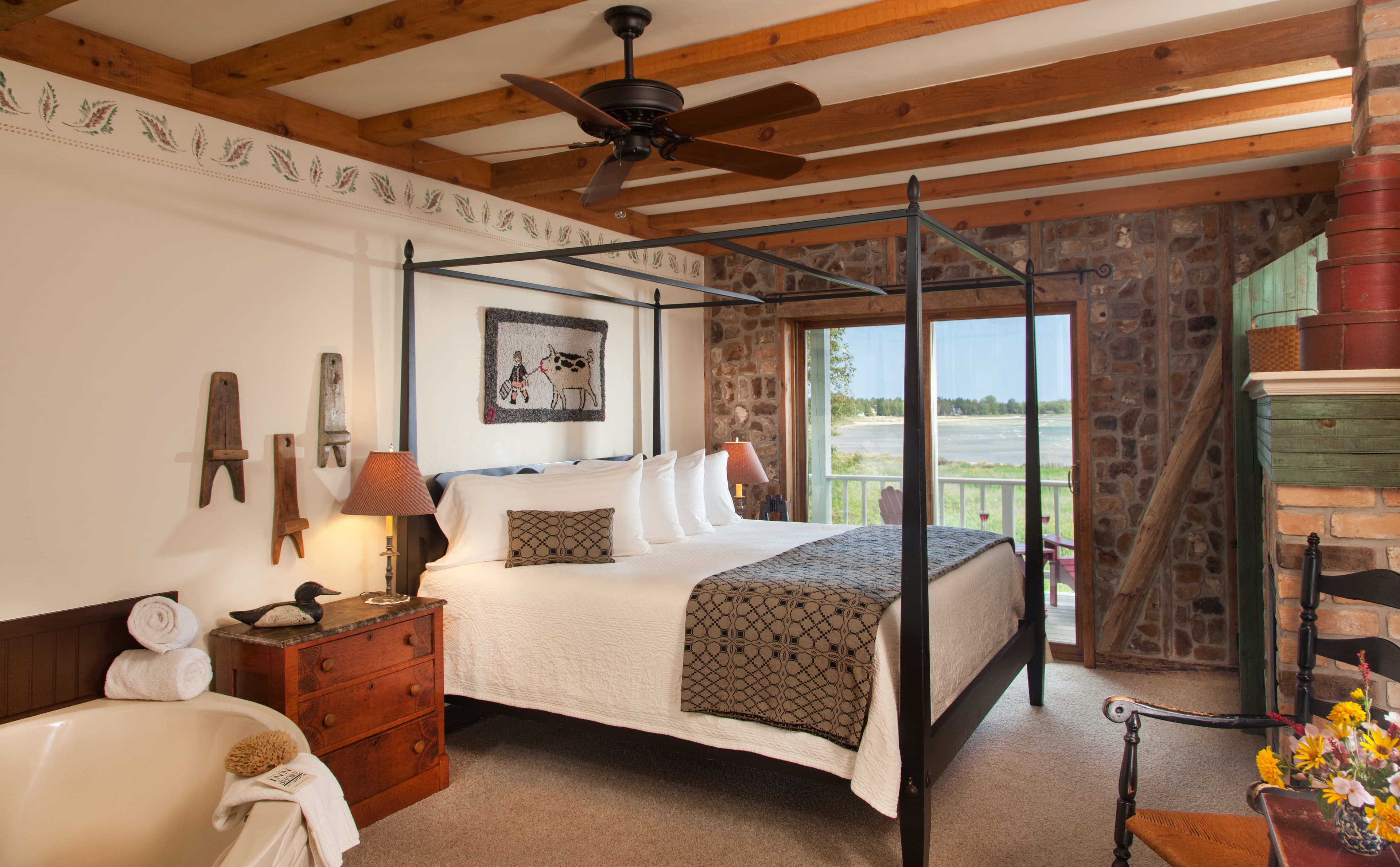 A spacious bedroom featuring a black metal canopy bed with white linens and a patterned throw, set against an exposed stone wall with a sliding glass door that leads to a balcony overlooking Lake Michigan.
