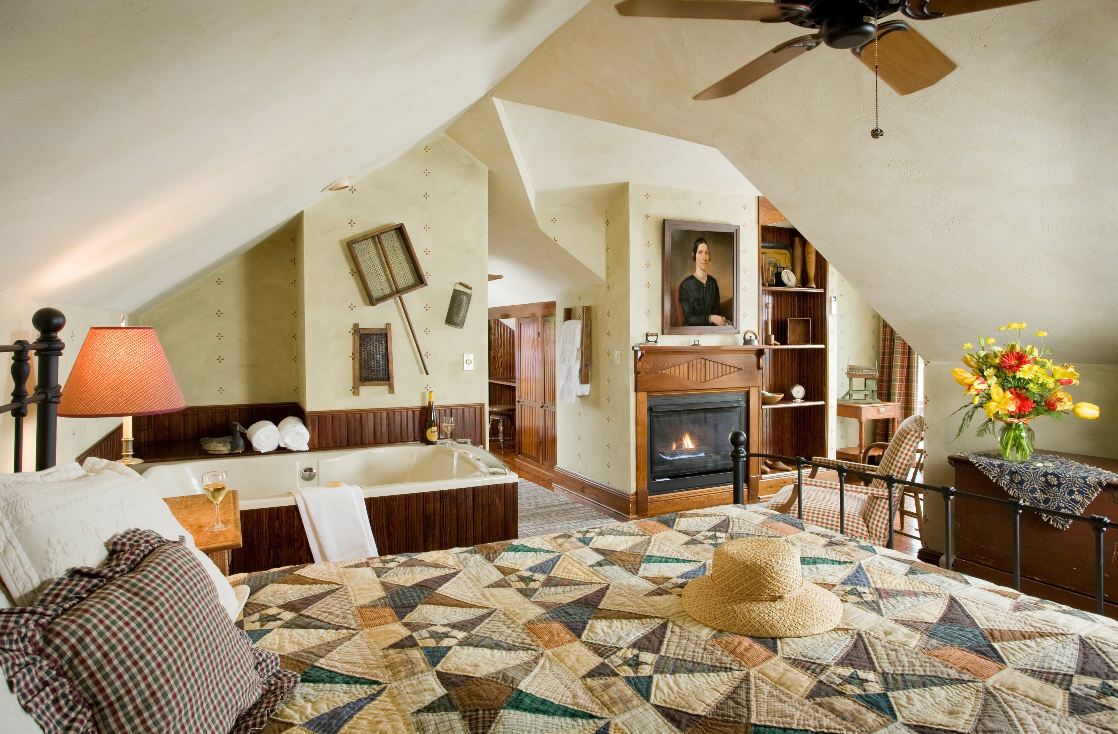 A cozy attic-style bedroom featuring a bed with a geometric patterned quilt, a whirlpool tub set against wood paneling, a lit fireplace under a traditional portrait, and slanted ceilings that create an intimate atmosphere.