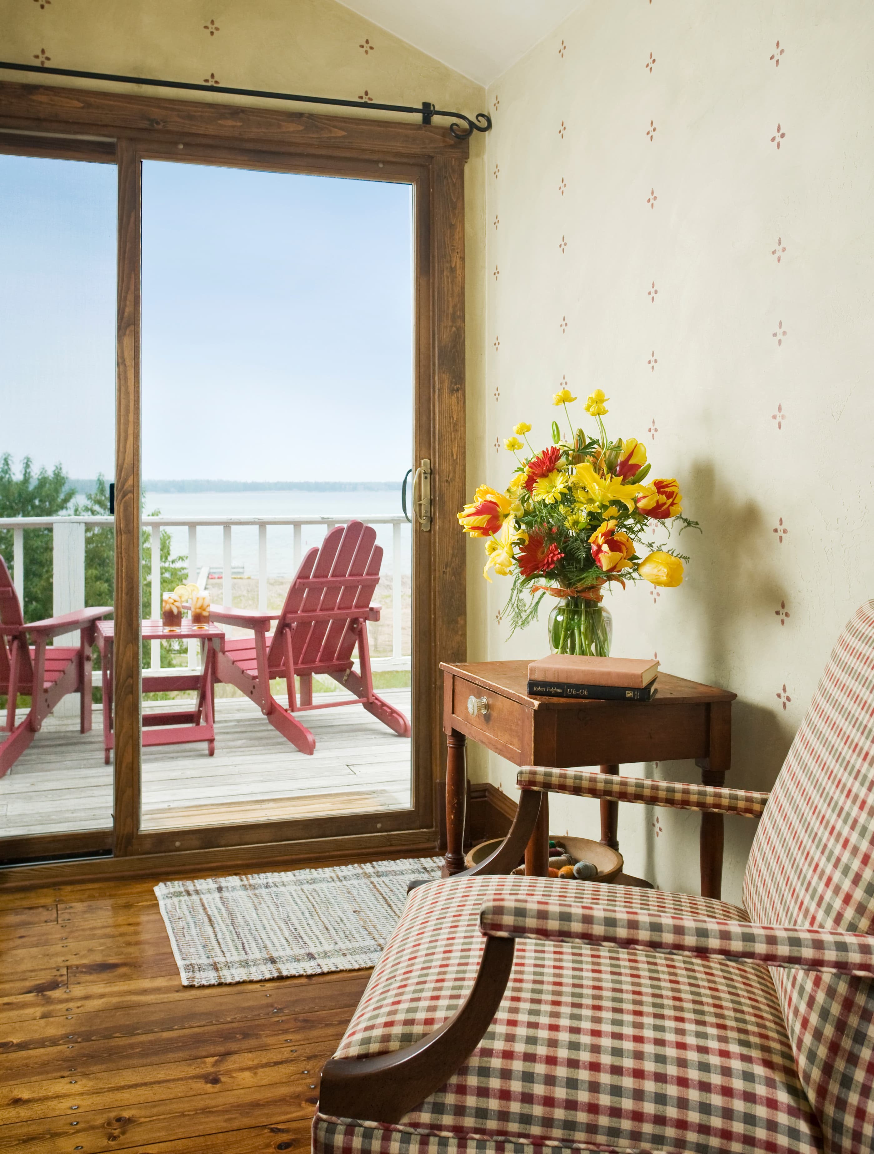 A cozy room interior featuring a red and white checkered armchair and a wooden side table with a vibrant bouquet of yellow and red flowers, situated next to a sliding glass door that leads to a balcony with red Adirondack chairs and a view of the water.