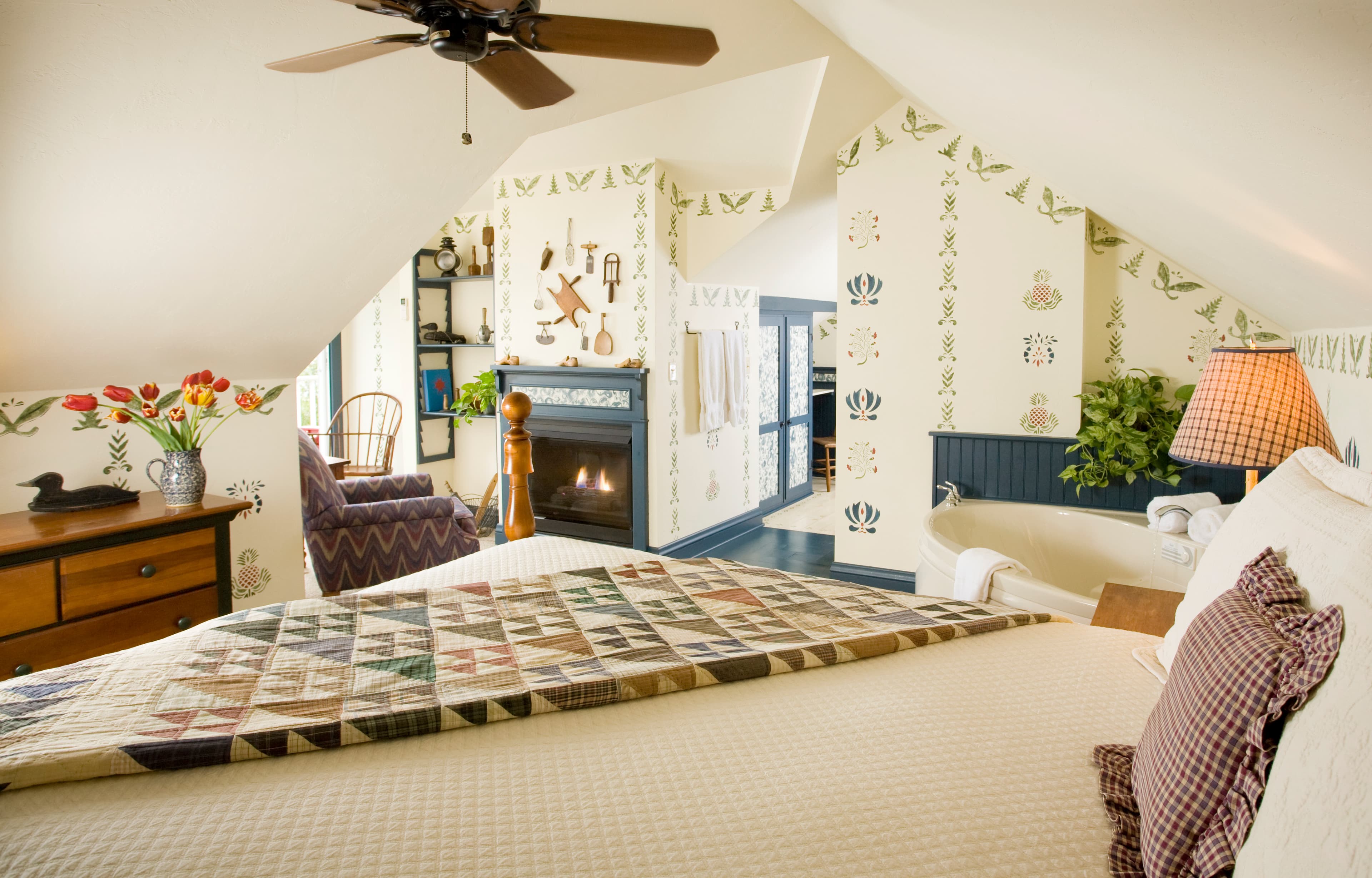 A cozy bedroom featuring a bed with a colorful geometric quilt, a lit fireplace with a blue mantel, a white whirlpool tub in the corner, and walls decorated with stenciled patterns.