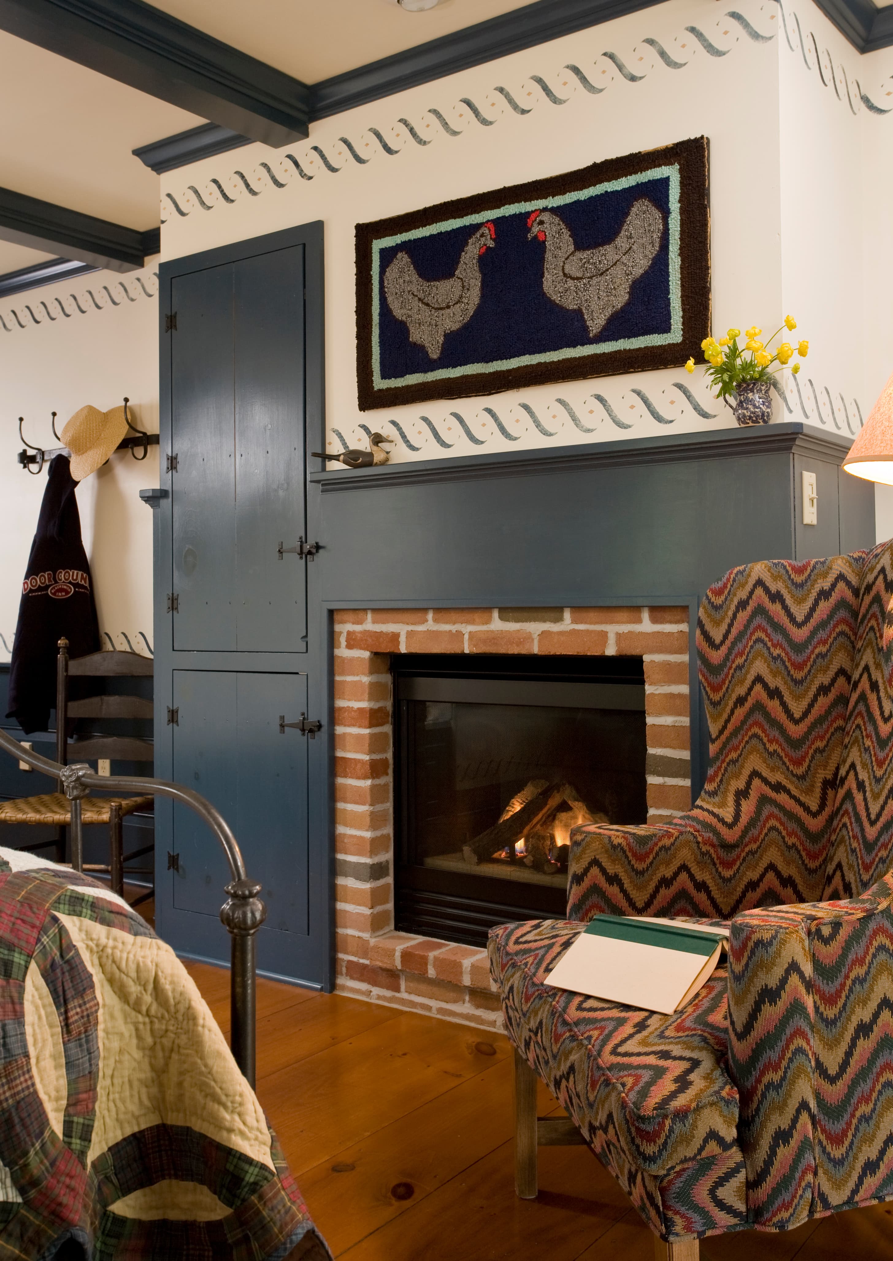 A cozy bedroom corner featuring a brick fireplace with a dark blue mantel, a patterned wingback chair with a zigzag design, and a piece of folk art depicting two chickens hanging on the wall above.