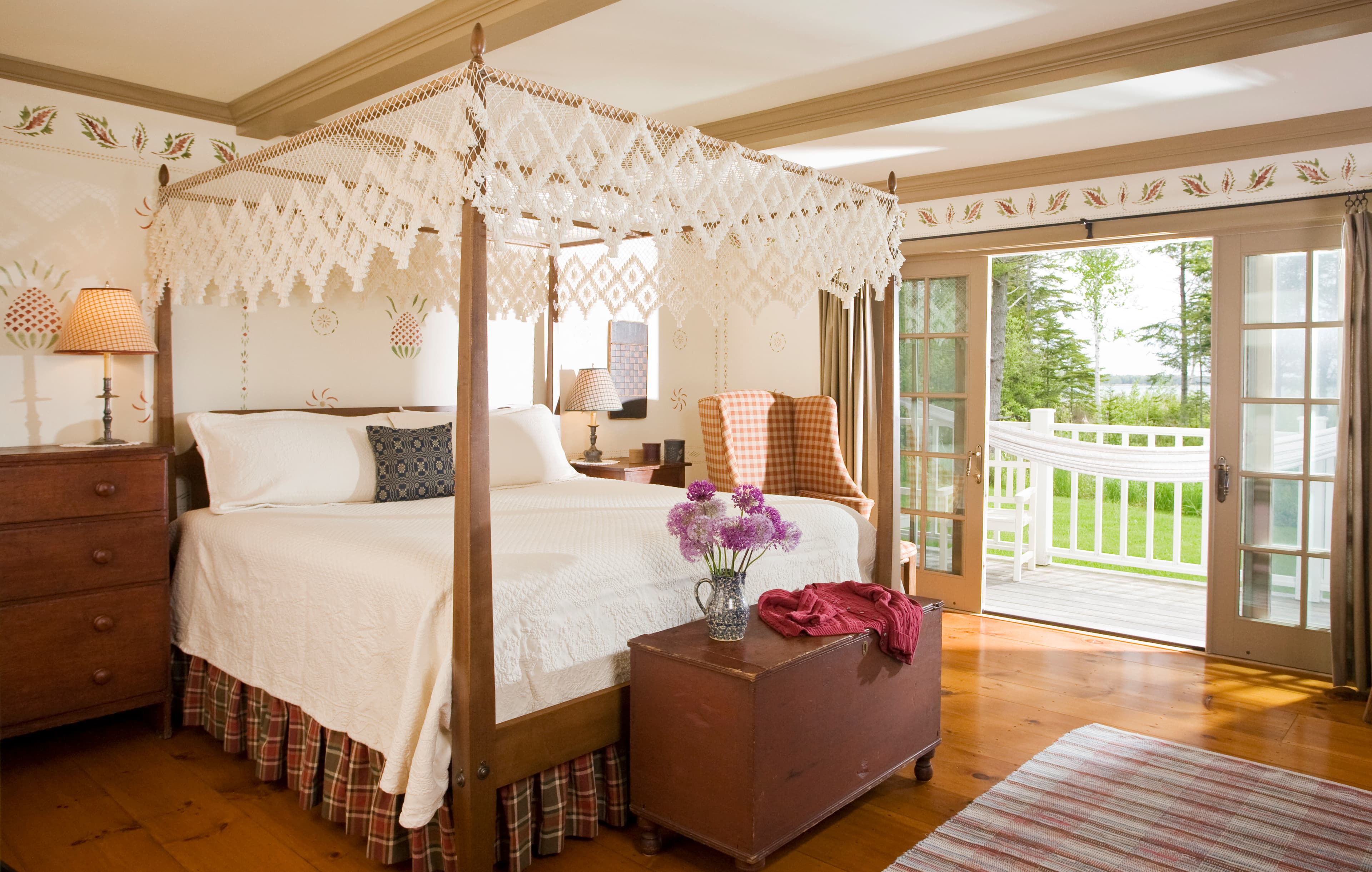 A spacious bedroom featuring a wooden four-poster bed with a white macramé canopy, a chest at the foot of the bed with purple flowers, and French doors leading to a balcony with a view of greenery and water.