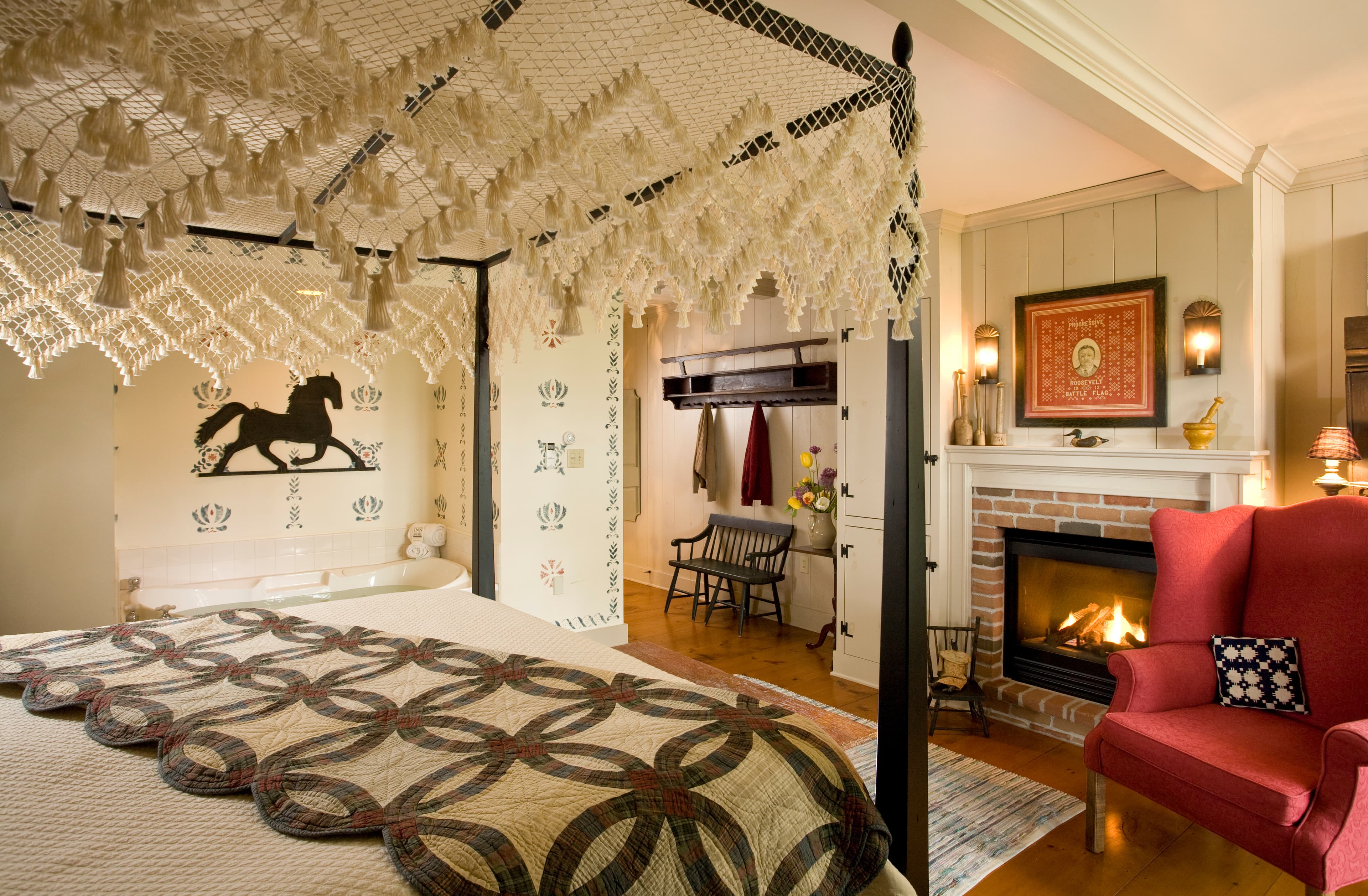 A spacious bedroom featuring a black canopy bed with a lace canopy, a brick fireplace with a white mantel, and a red armchair in the foreground.