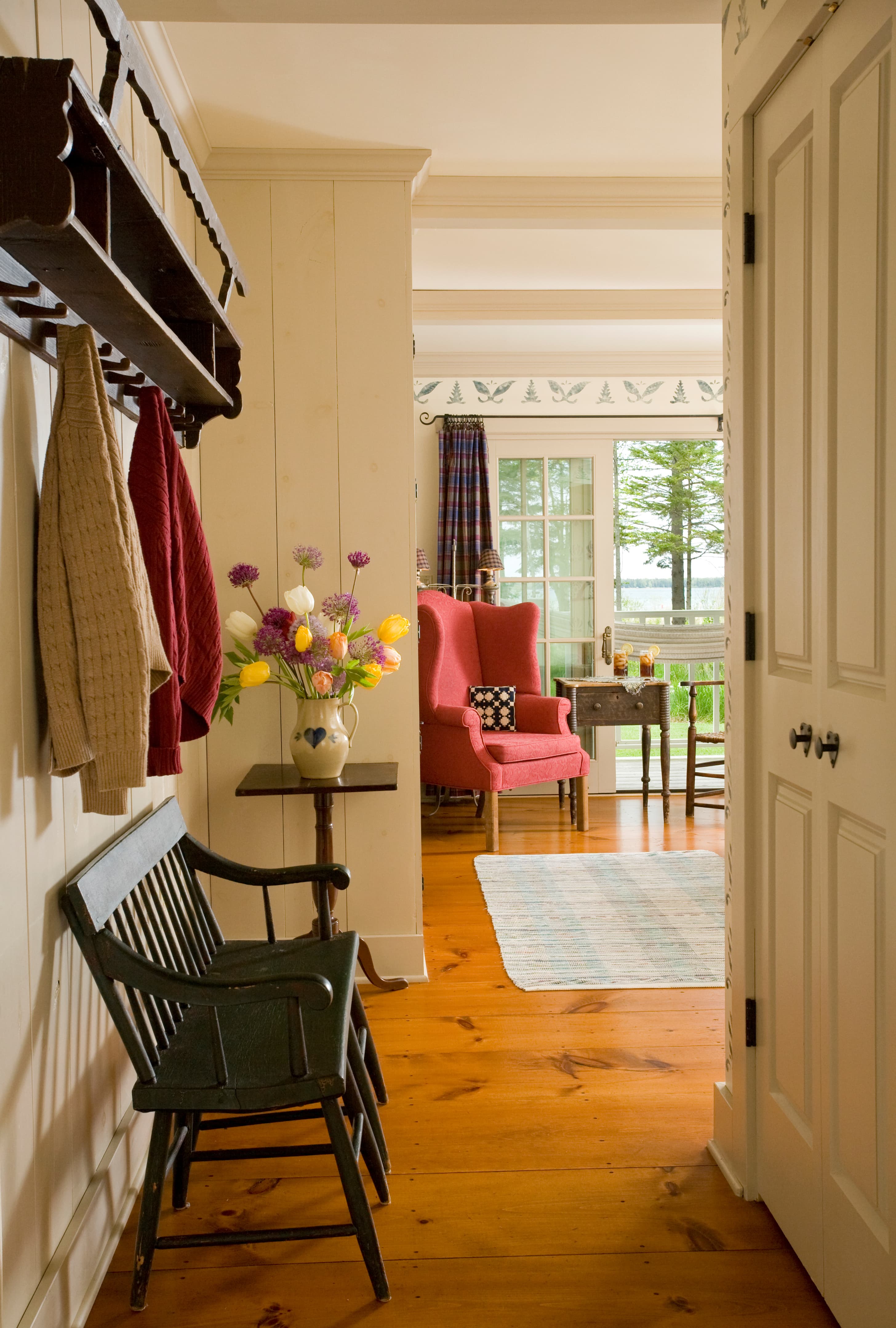 A hallway with polished wood floors featuring a dark green wooden bench and a wall-mounted coat rack with hanging sweaters, leading into a bright room with a red wingback chair and glass doors opening to a scenic outdoor view.