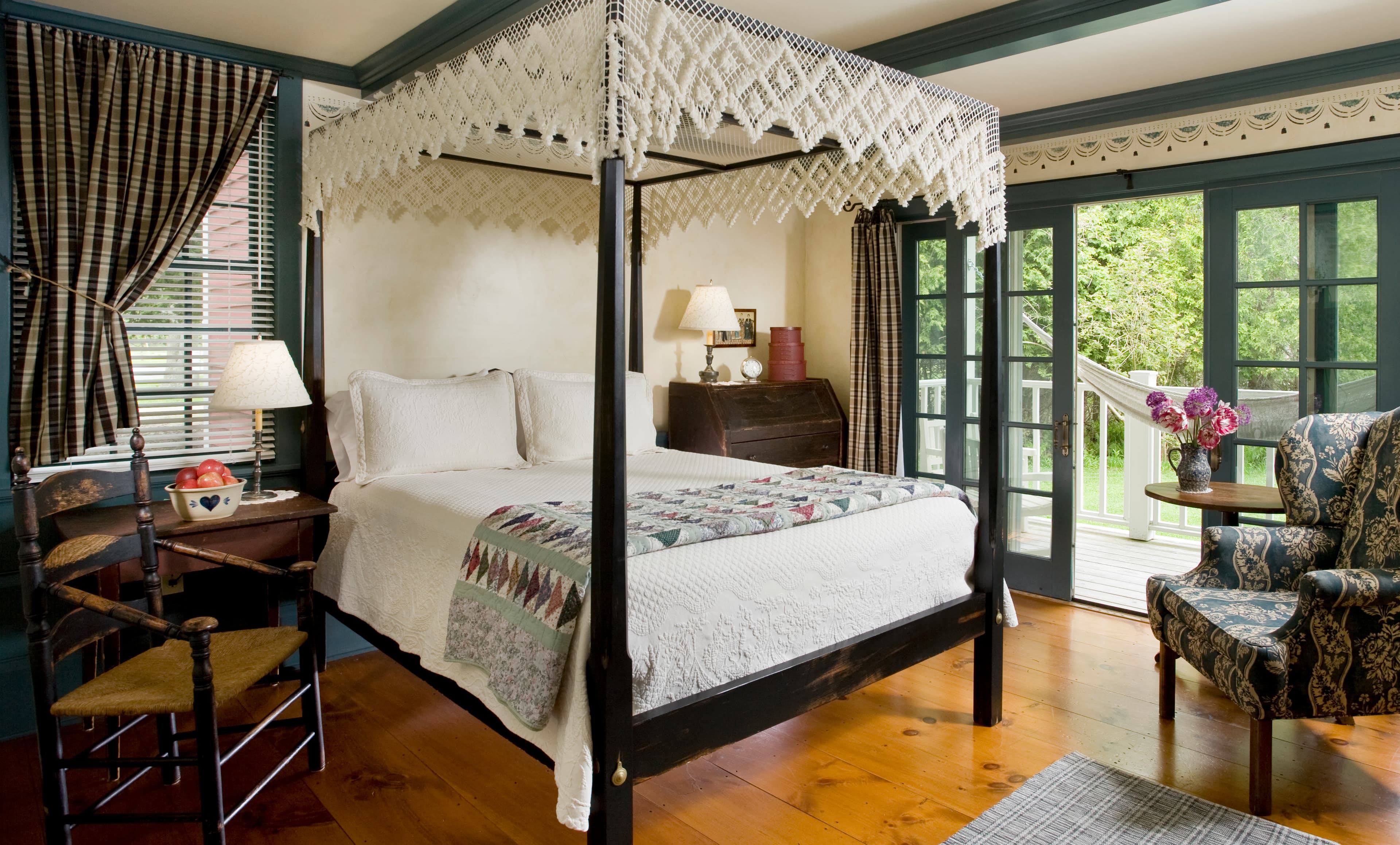 A bedroom with dark blue trim and ceiling beams, featuring a black canopy bed with a white lace canopy and a patterned throw, a patterned wingback chair, and glass doors leading to a balcony with a white hammock.
