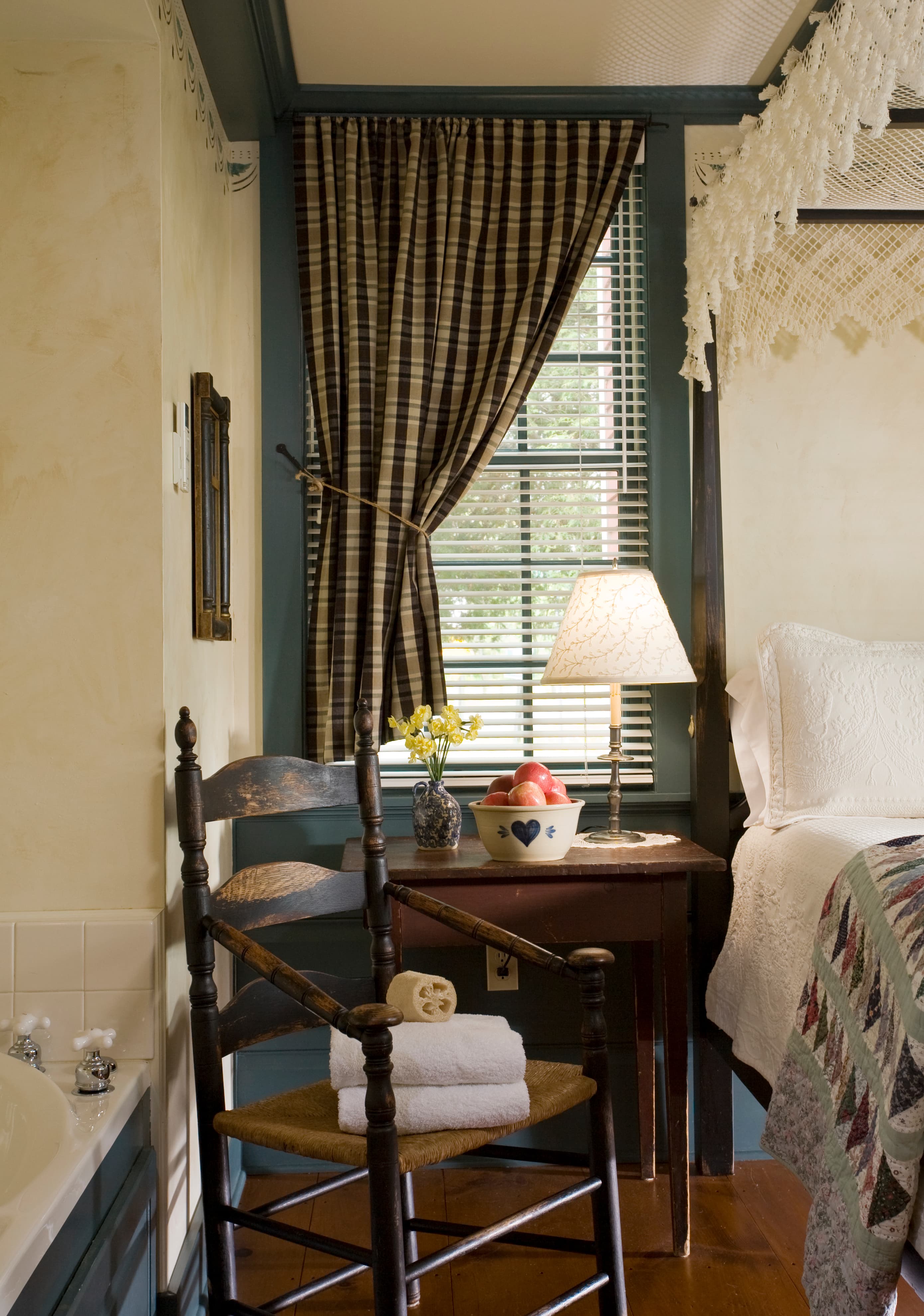 A bedroom with blue-painted ceiling beams and trim, featuring a bed with a double wedding ring patterned quilt, a patterned wingback chair, and french doors that open to a balcony with a white hammock and a view of trees.