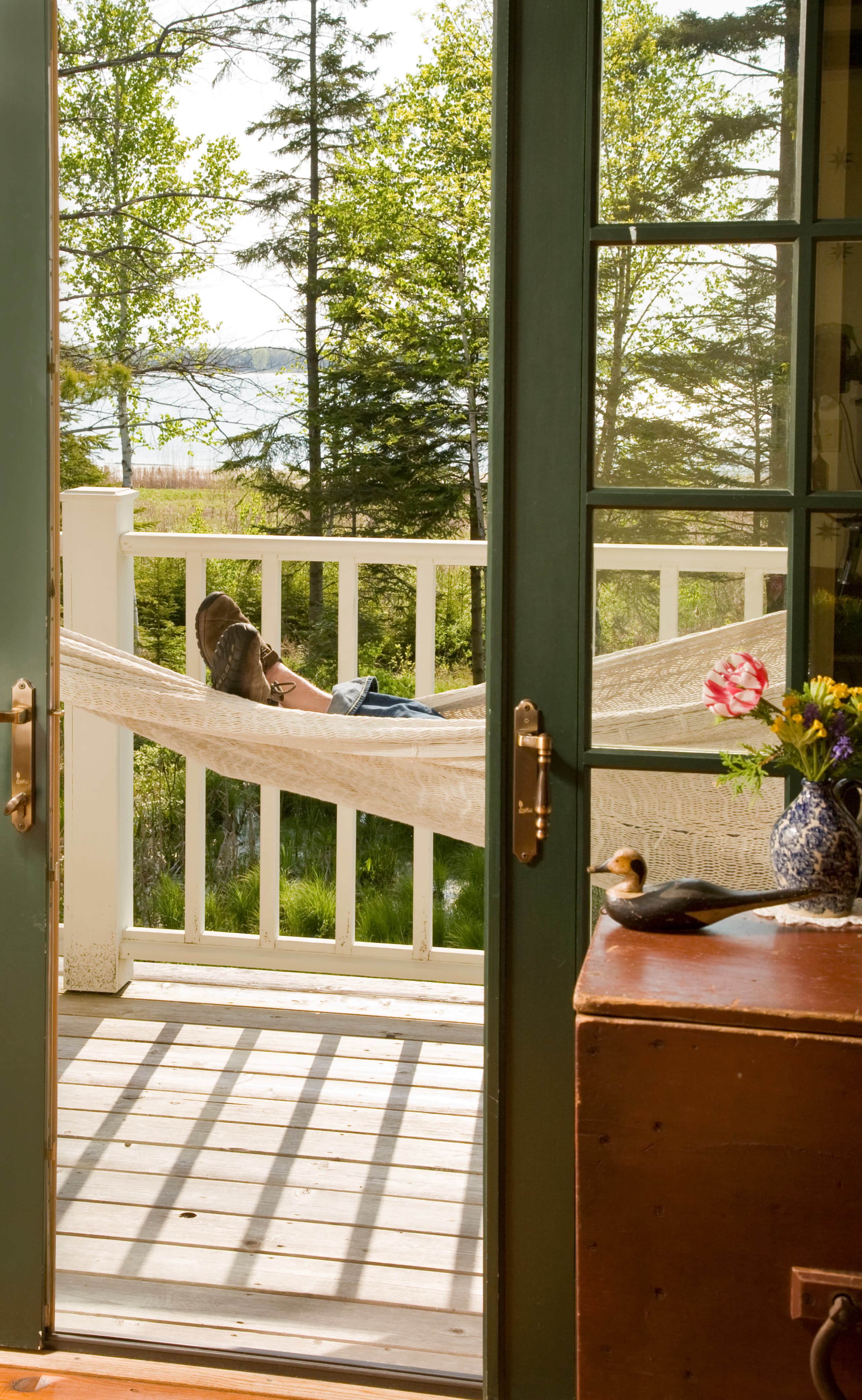 A close-up view of a person's feet resting in a white rope hammock on a balcony, with a white railing and a scenic background of green trees and a body of water.