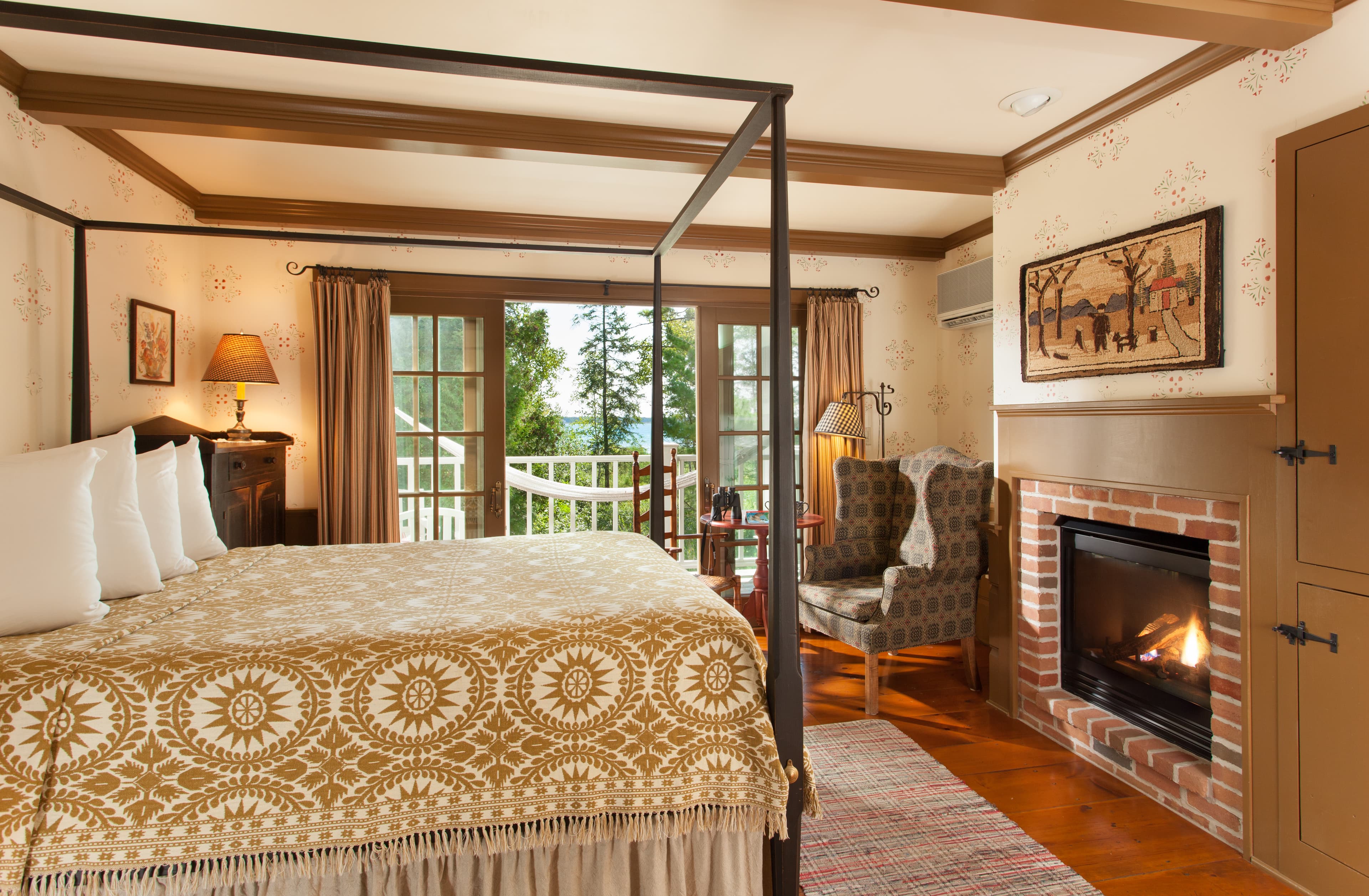 A spacious bedroom featuring a black metal canopy bed with a tan and white patterned quilt, a brick fireplace with a roaring fire, and glass doors leading to a balcony with a white hammock and a view of lush greenery.