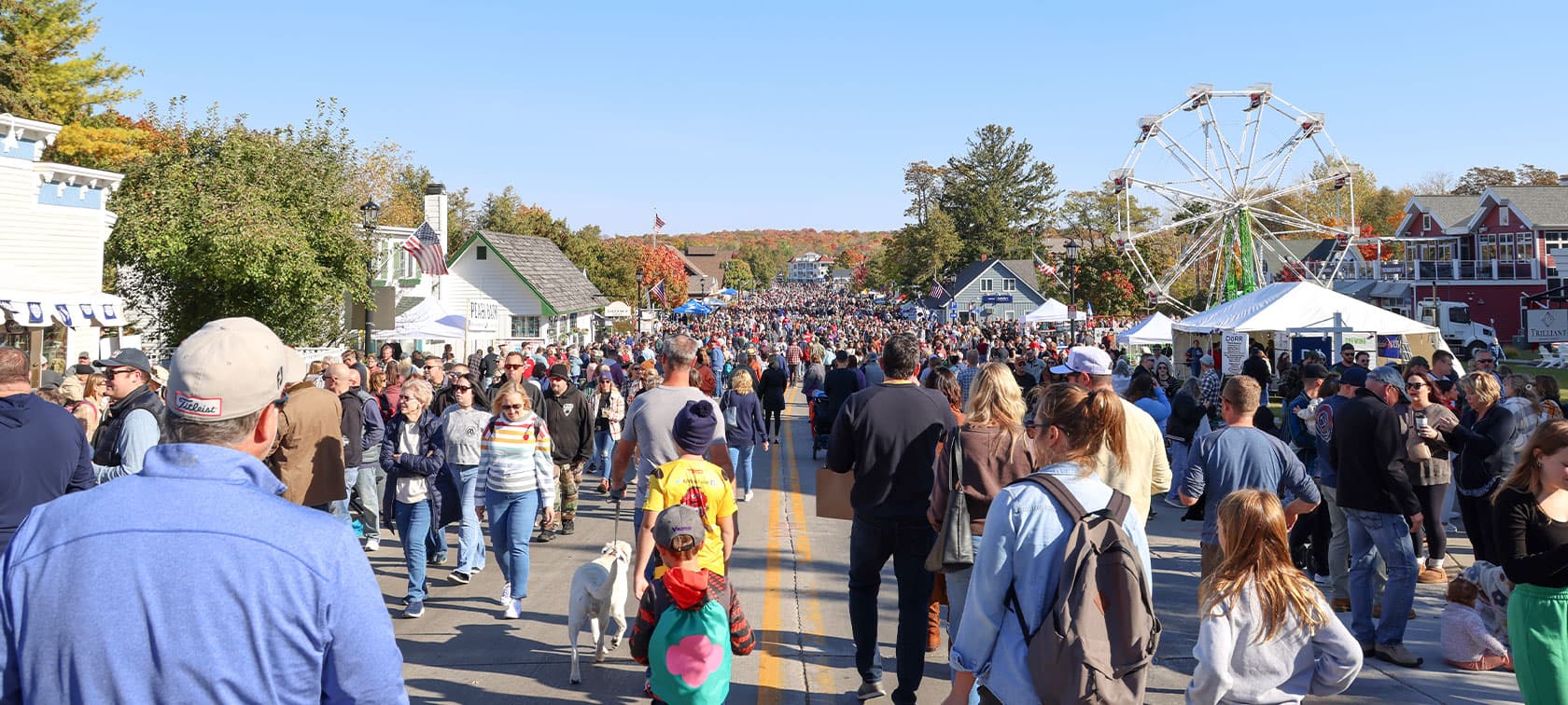 A large crowd gathers on a sunny street lined with tents and a ferris wheel in the background. A large crowd gathers on a sunny street lined with tents and a ferris wheel in the background.
