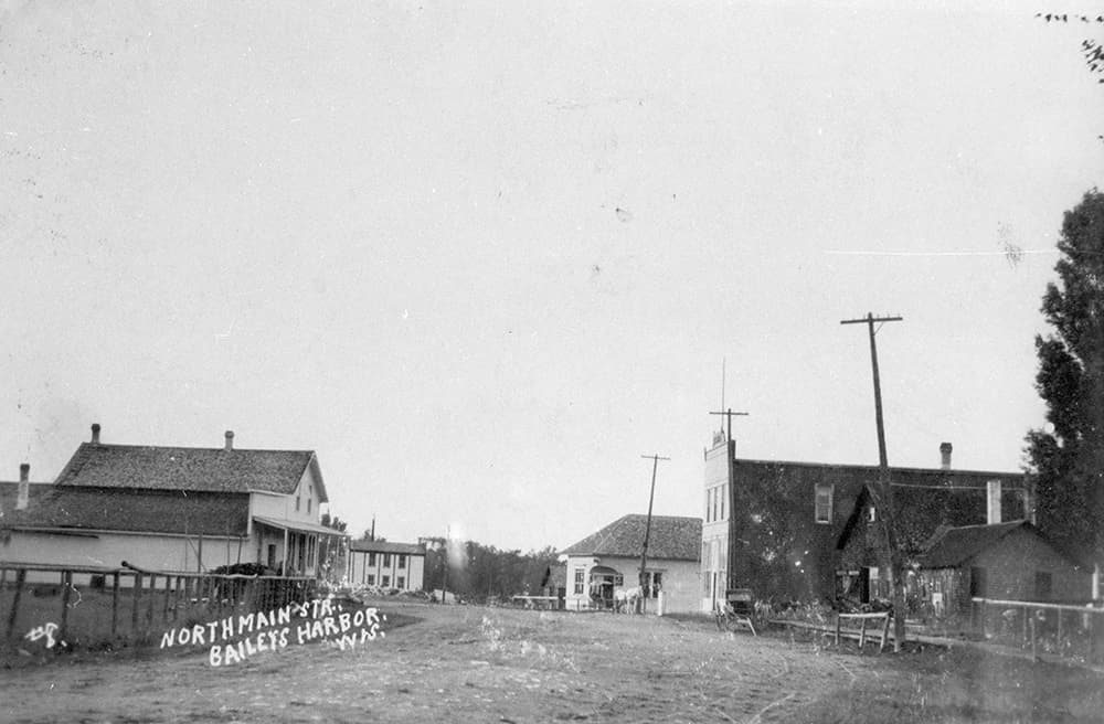 Historic black-and-white photo of North Main Street in Bailey's Harbor, showcasing early buildings and telephone poles.