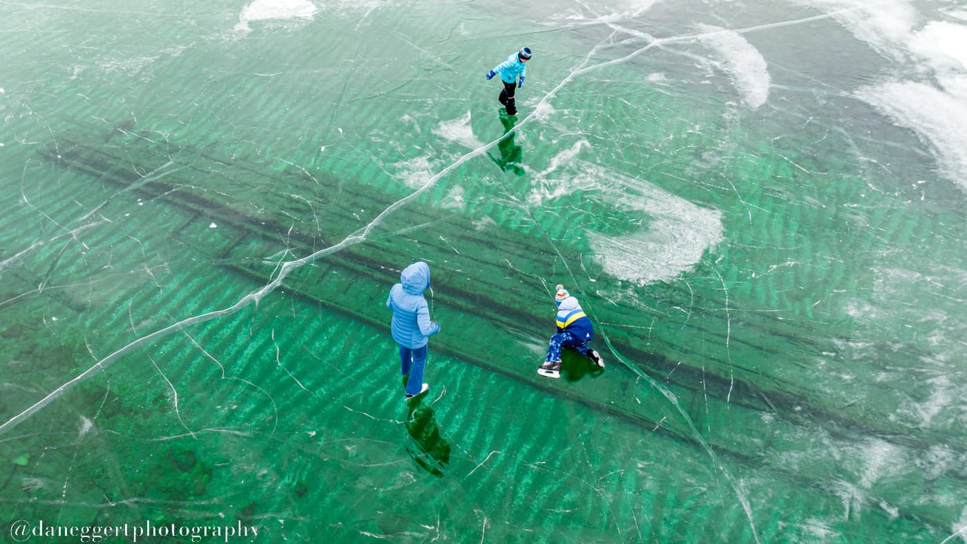Children play on a frozen lake, exploring the clear ice above a submerged boat.