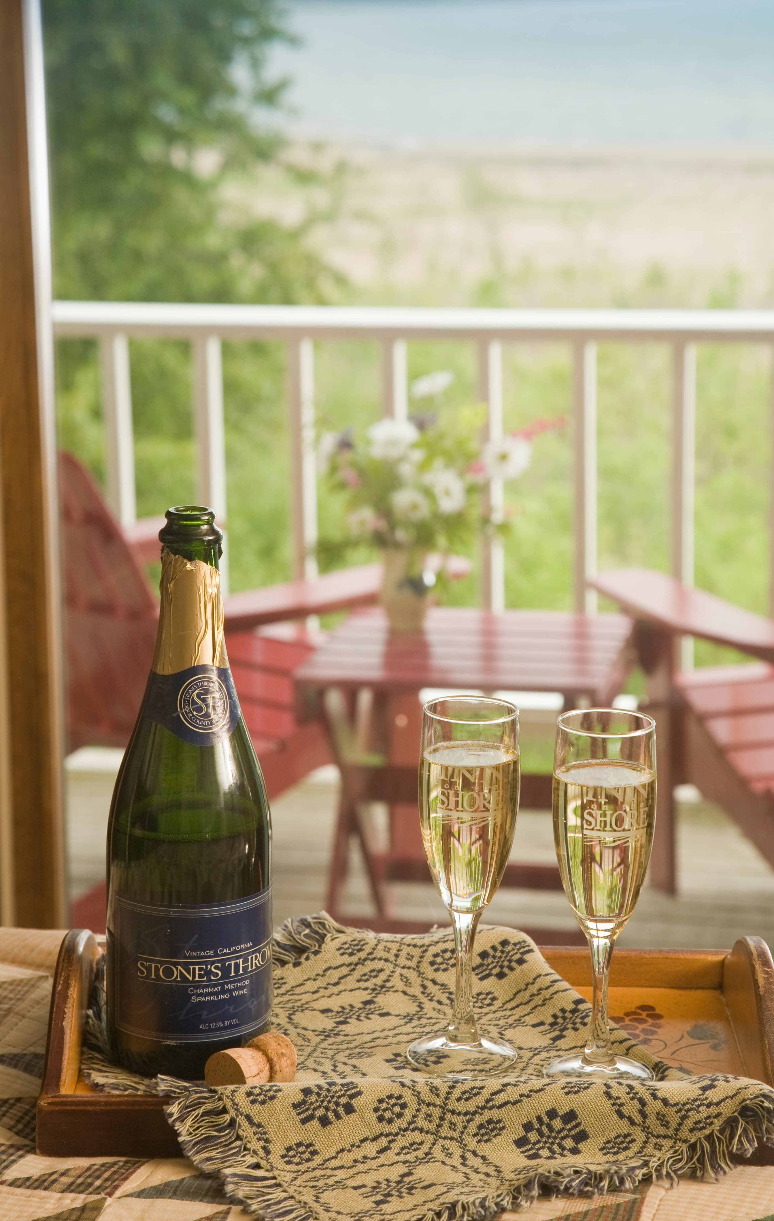 A bottle of sparkling wine and two filled glasses sit on a tray with a scenic view of a deck and garden.
