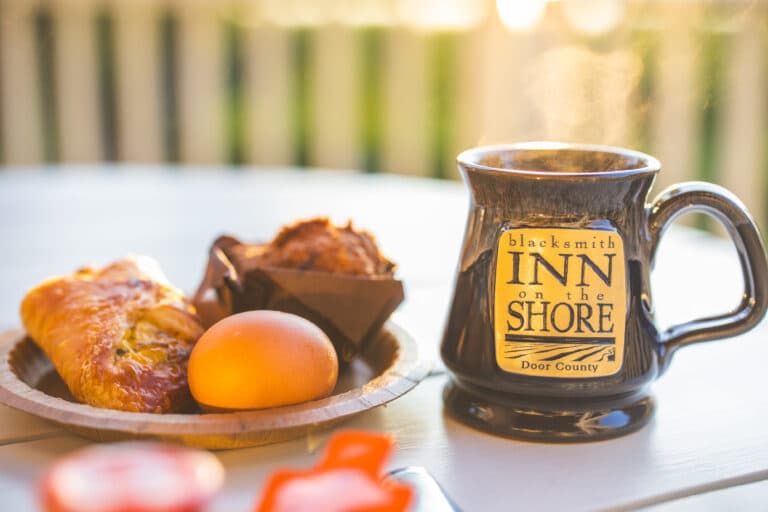 A steaming coffee mug beside a breakfast plate with an egg, muffin, and pastry.