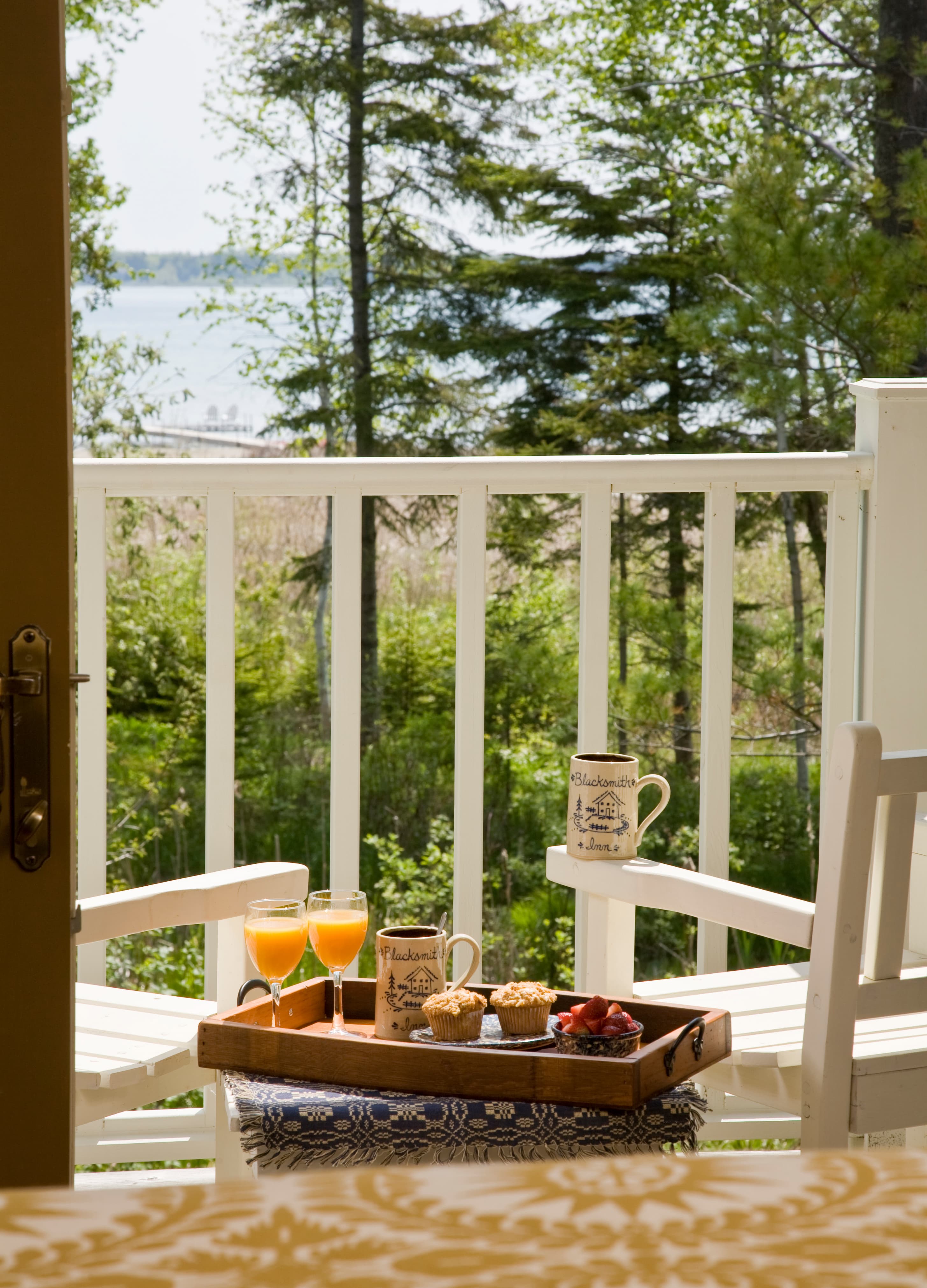 A sunlit balcony with white wooden railings and chairs overlooking a body of water and lush green trees, featuring a wooden breakfast tray with two glasses of orange juice, muffins, and mugs.