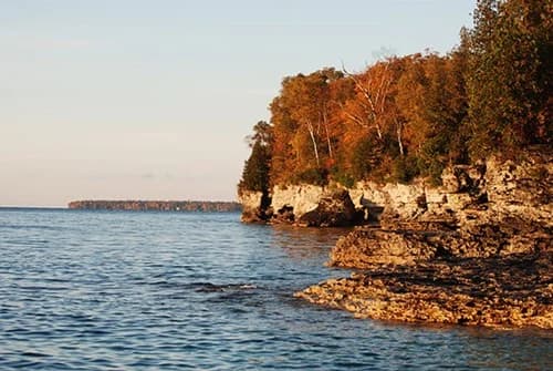 Rocky shoreline with autumn-colored trees and calm water under a clear sky.