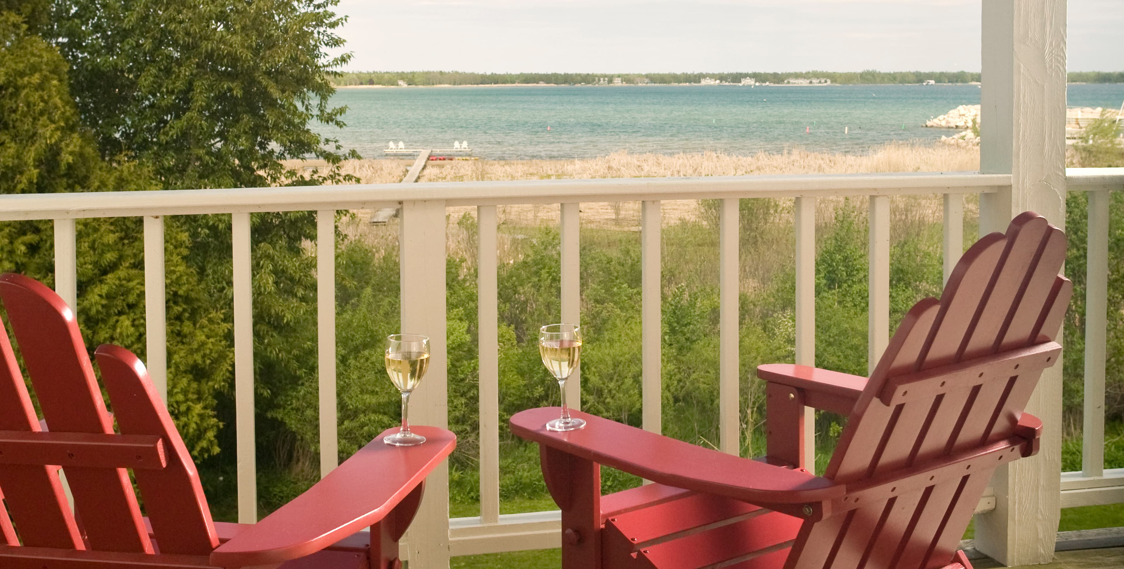 Two red adirondack chairs with glasses of wine overlook a serene waterfront scene.