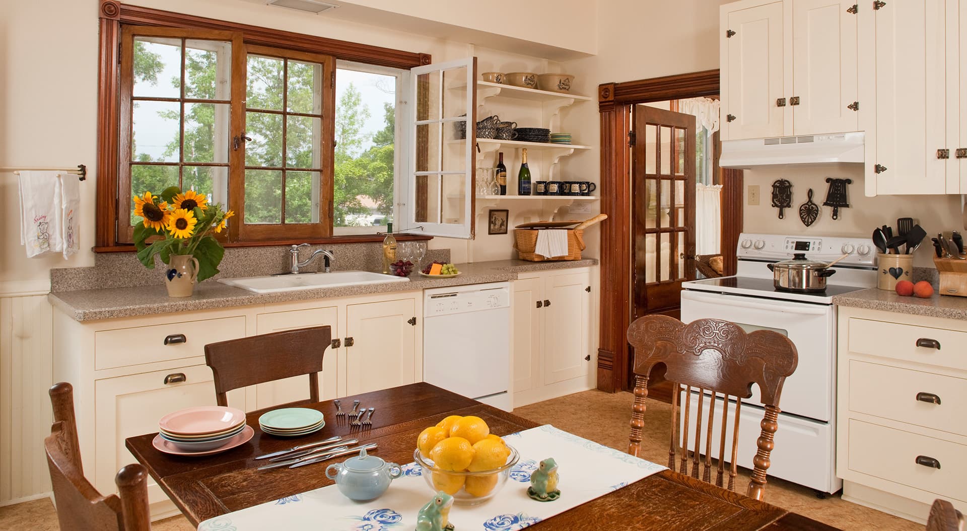 A bright, cozy kitchen featuring white cabinets, a wooden table with dishes, and a window view of greenery.