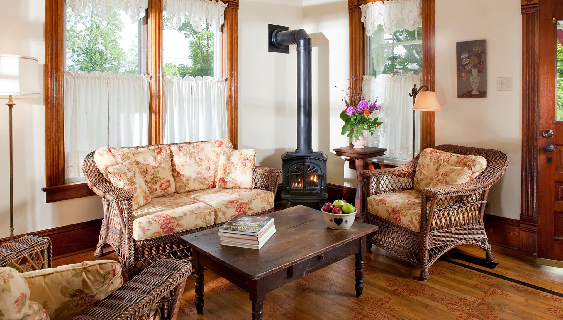 A cozy living room with floral-patterned wicker furniture, a wooden coffee table, and a small wood stove.