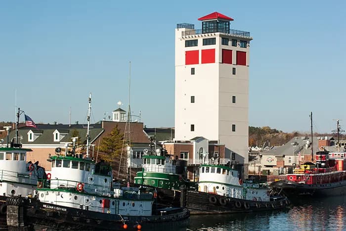 Tugboats lined up at a harbor next to a tall, white tower with a red roof.