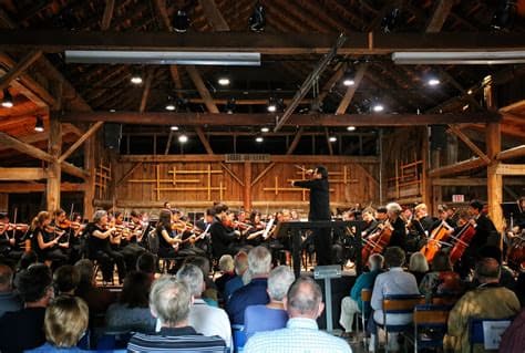 A conductor leads an orchestra performing in a rustic barn setting, while an audience watches.