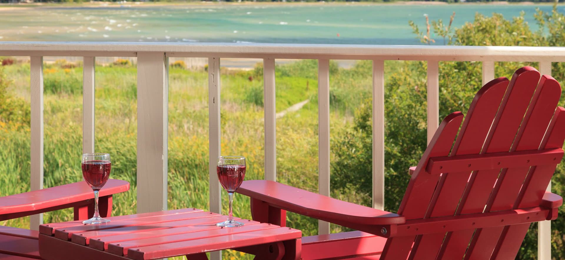Two red chairs and a small table with glasses of wine overlook a serene, green landscape and water.
