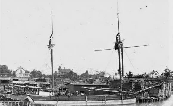 A historic black-and-white photo of a sailboat docked by a pier with wooden buildings in the background.