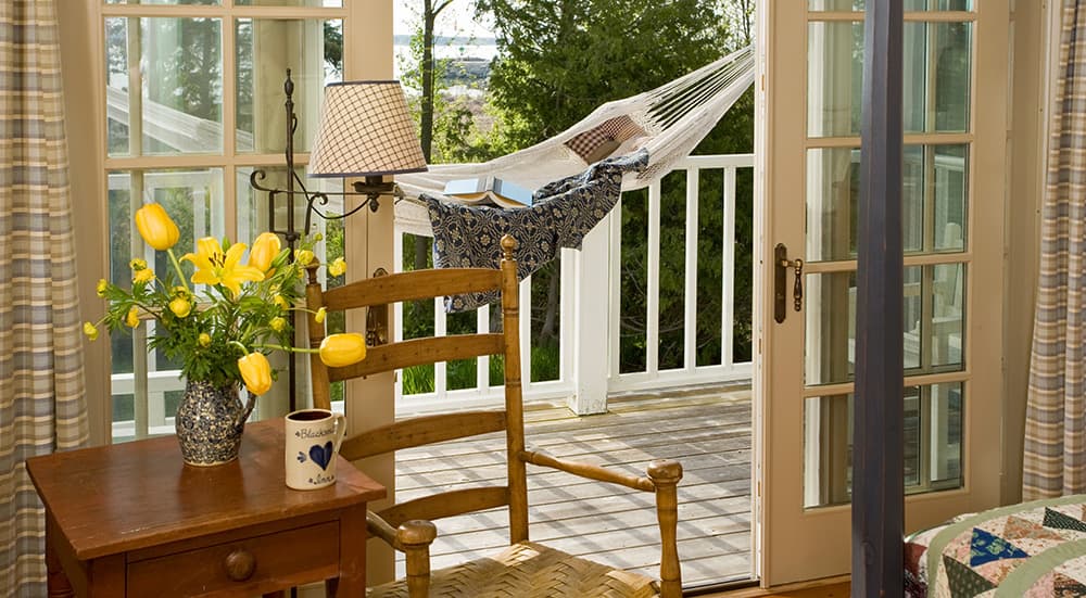 A cozy room with a wooden chair, a table with flowers, and a hammock visible through glass doors leading to a balcony.