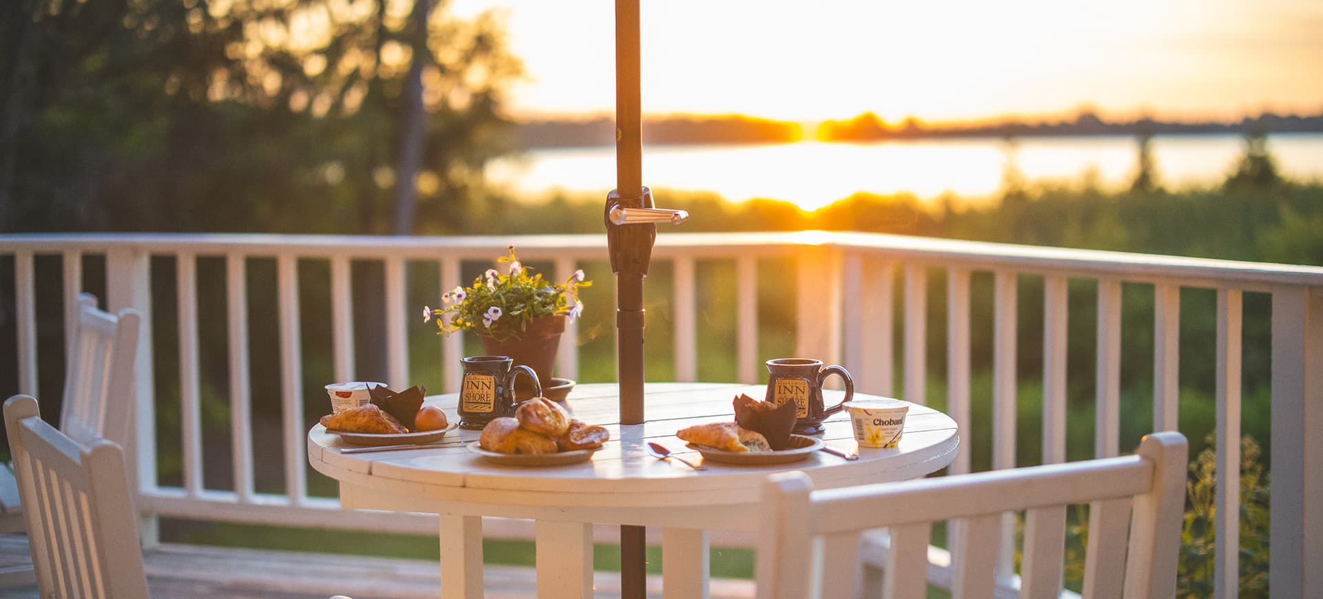 A table set for breakfast on a balcony overlooks a serene sunset by the water.