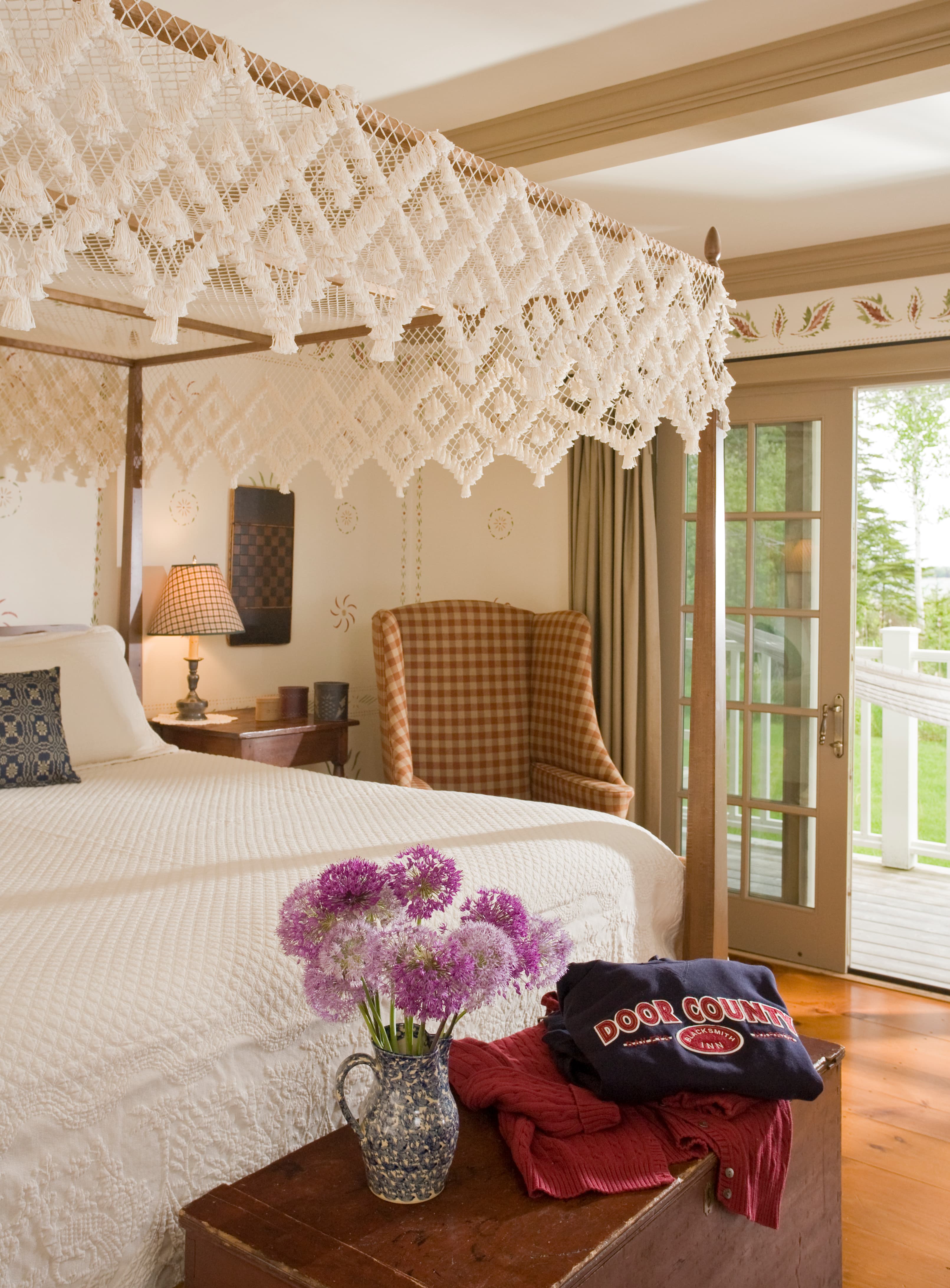 A cozy bedroom featuring a canopy bed, a checkered armchair, and a vase of purple flowers on a wooden chest.