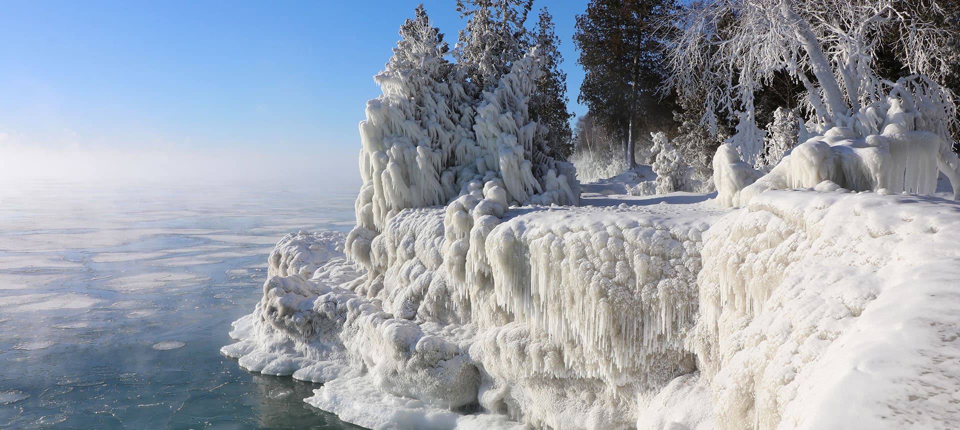 Frost-covered cliffs and trees overlook a misty, ice-covered lake.
